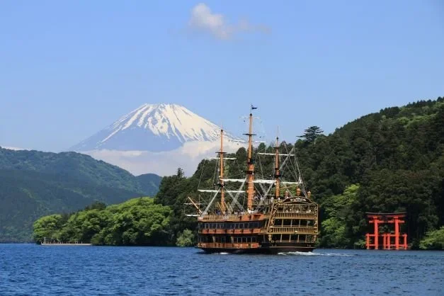 Snow covered Mount Fuji in the background with a lake with a tori gate and a pirate ship in the foreground.
