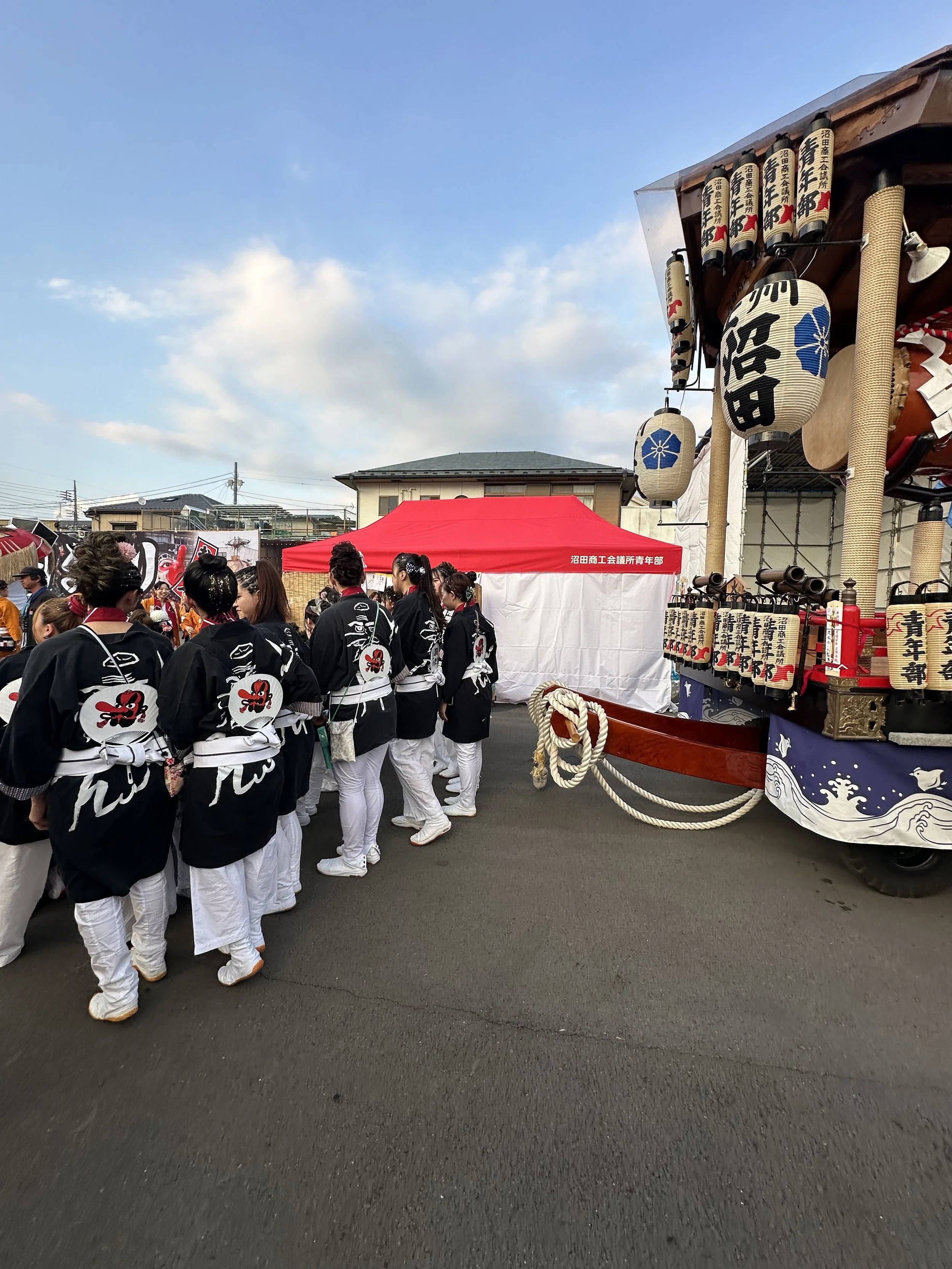 A group of people dressed in traditional Japanese festival clothing, gathered around a float decorated with lanterns and ornate details, during a cultural event outdoors on a clear day.