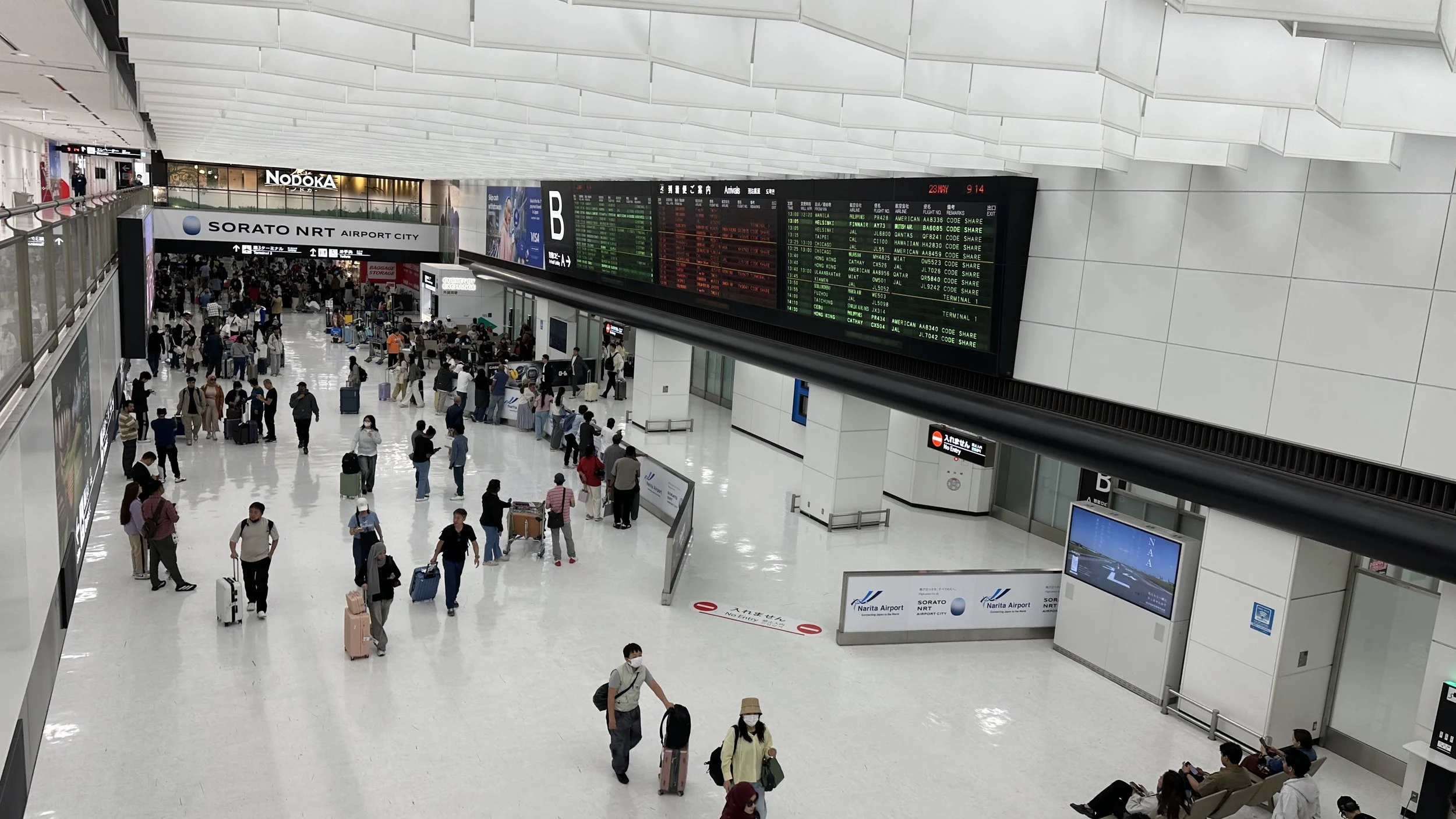 Interior of Narita Airport with travelers walking through the spacious departure hall, digital flight information display board showing flight details, and signs directing to various airport services.