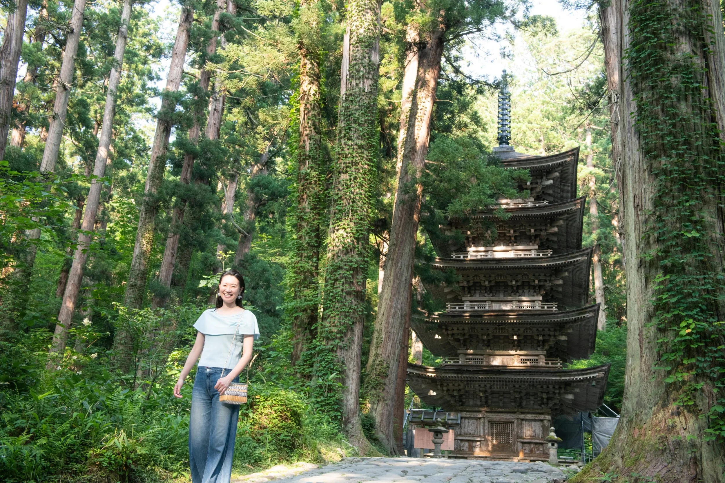5-story wooden Japanese Pagoda in a mountain surrounded by tall trees with a stone walk way in front and woman posing..