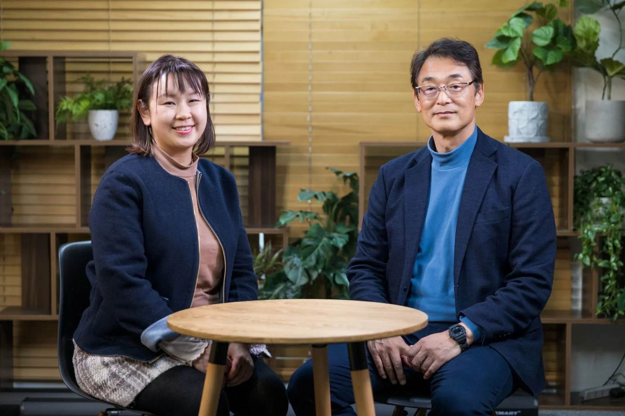 Two people sitting at a round wooden table in a room with wooden paneling and shelves with houseplants, smiling at the camera.