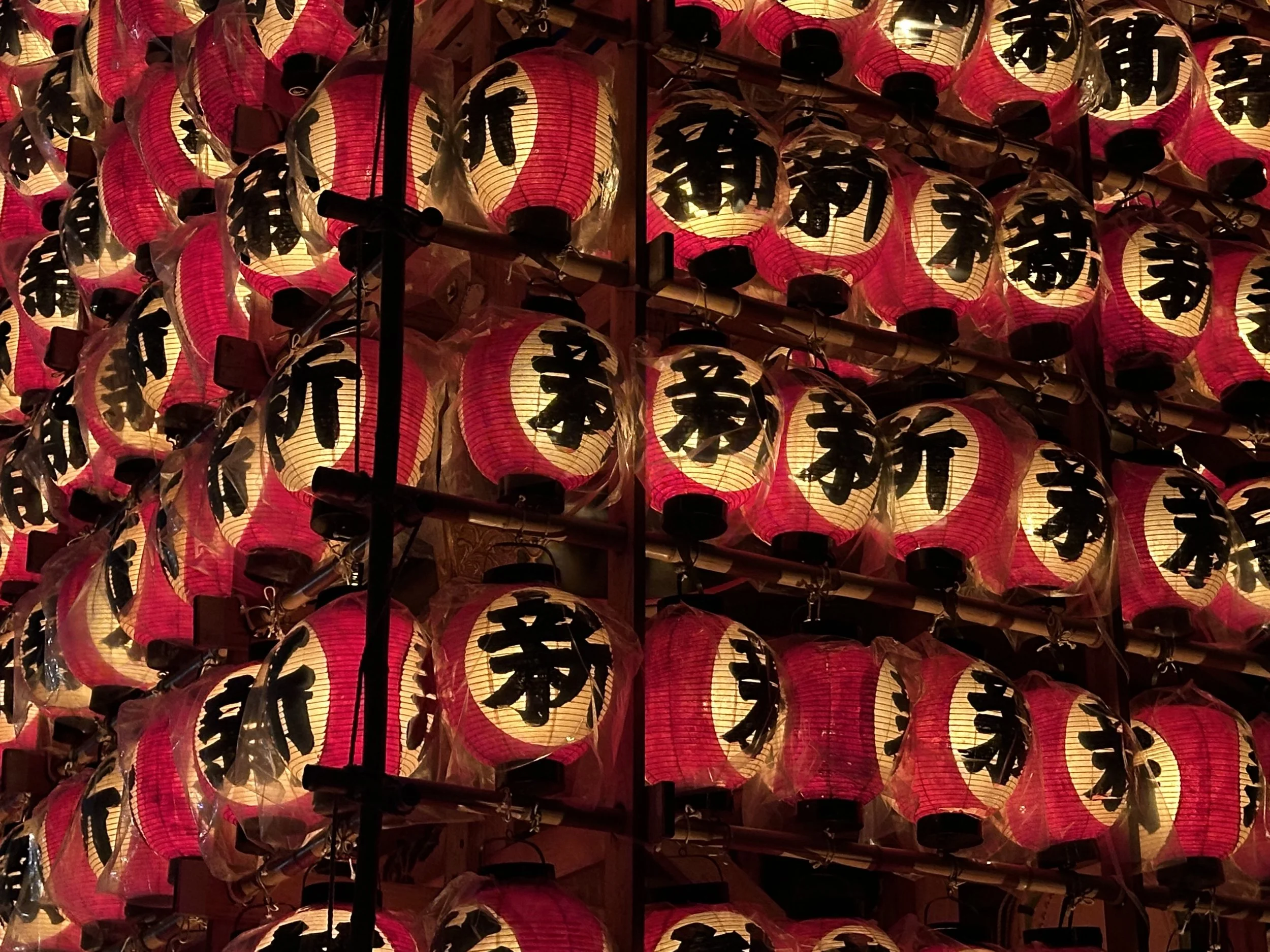 A large display of traditional Japanese paper lanterns, predominantly red and white, with black Japanese characters on each lantern, hanging closely together.