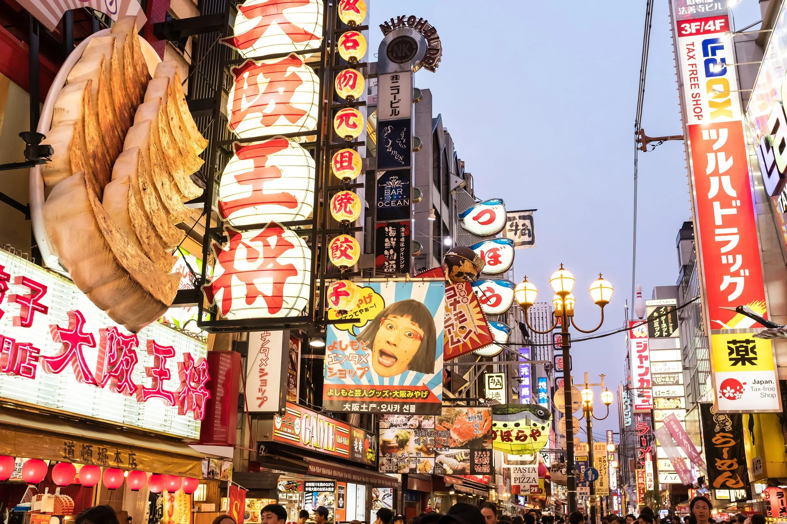 Busy Japanese street at dusk with shorter Showa-era buildings and vibrant night signage.