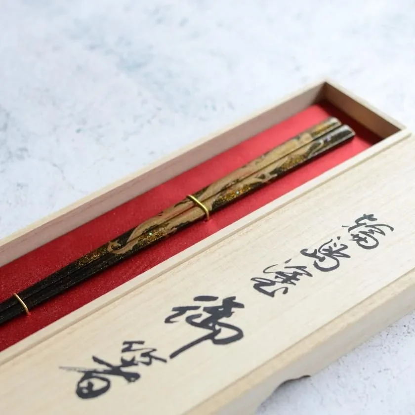 A set of golden and black chopsticks inside a wooden box with calligraphy letters.
