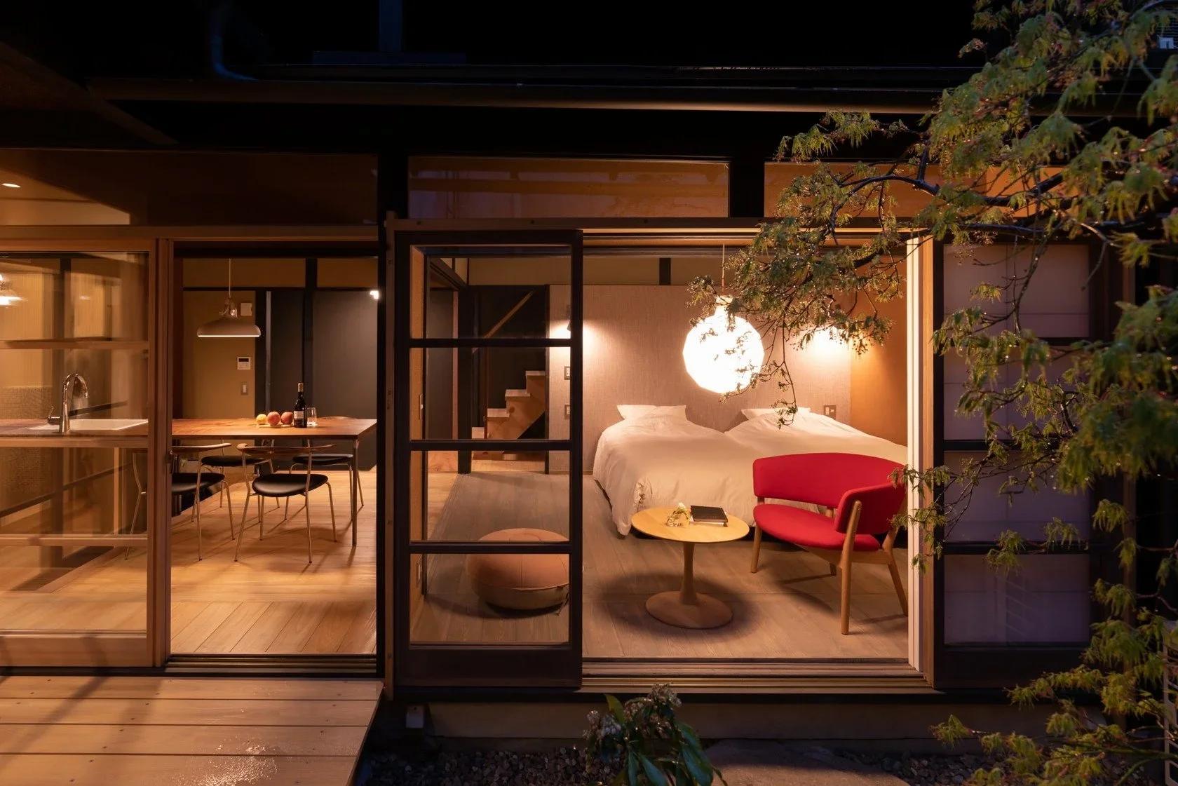 Modern Japanese home with kitchen and 4 chairs, bedroom with 2 single beds, and seating area with red chair, and a staircase with a tree in the foreground.