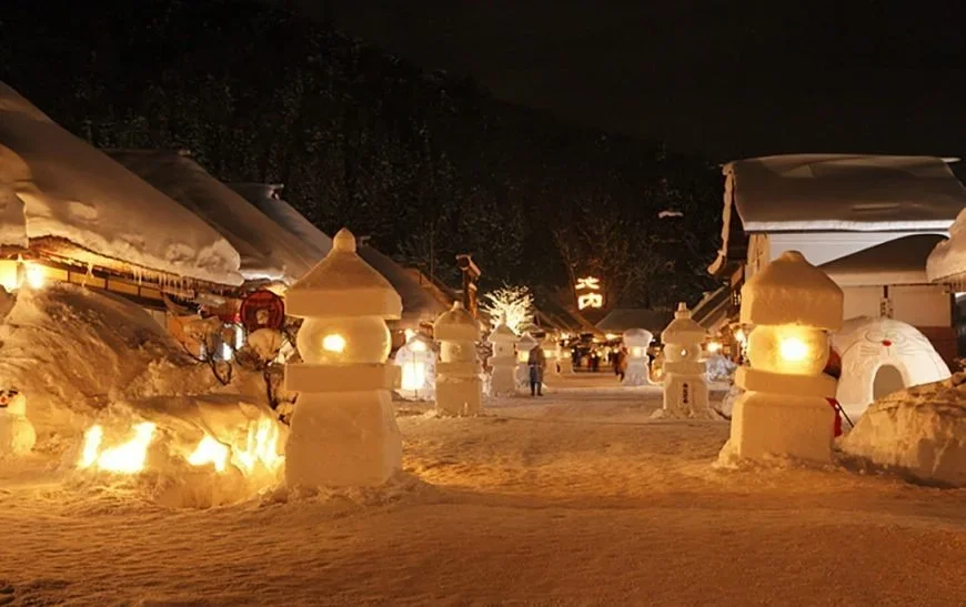 Lanterns made of snow that look like traditional stone lanterns pave a pathway among a street of traditional Japanese village homes and a flowing fire symbol of Japanese kanji in the background.