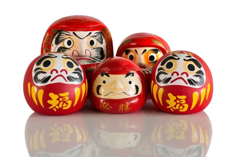 A variety of 5 round red dolls with yellow lines and Japanese Kanji characters painted on them.
