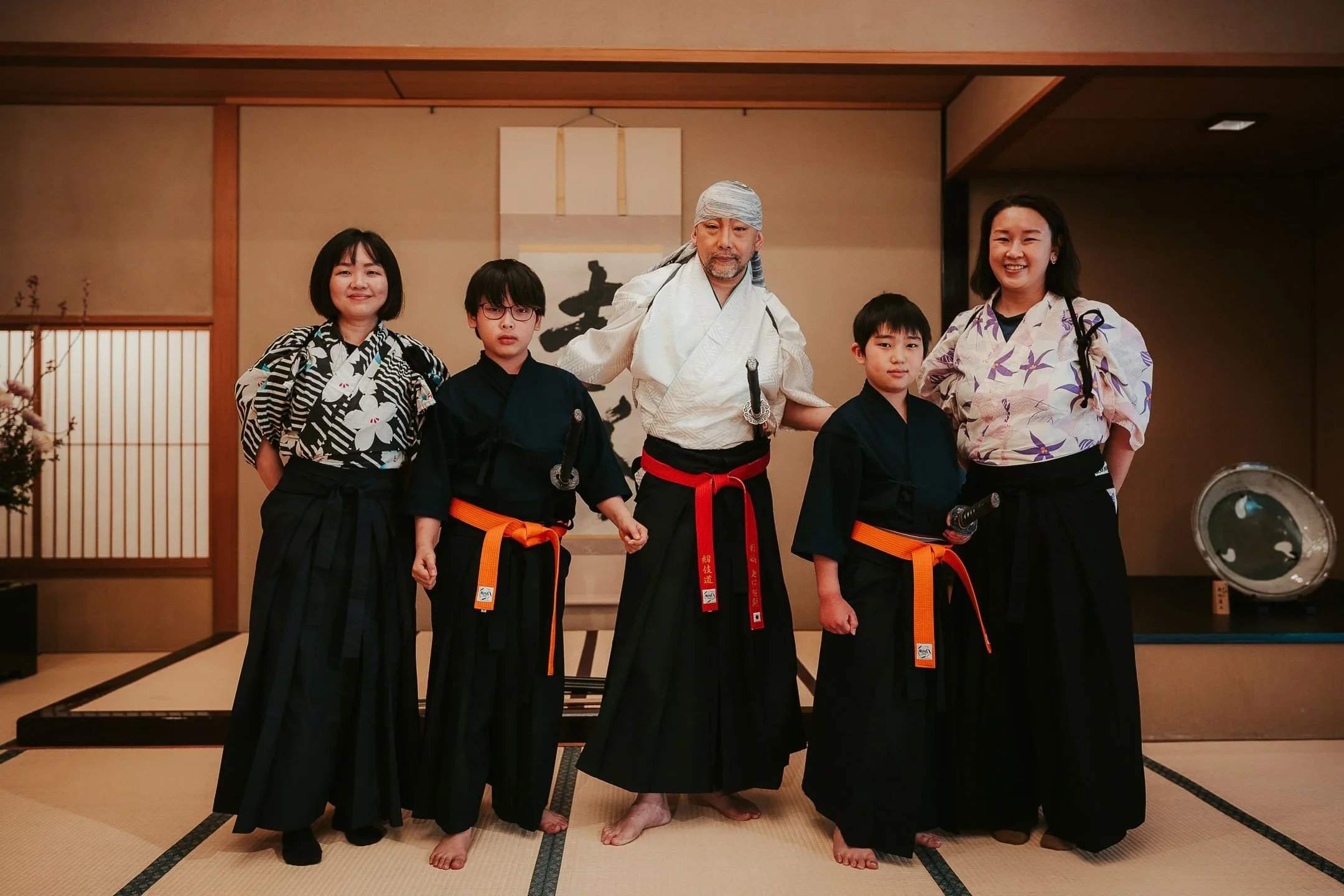5 member group including 2 kids, 1 man and two women dressed in a kimono and black hakama post in a line in a tatami mat Japanese room.