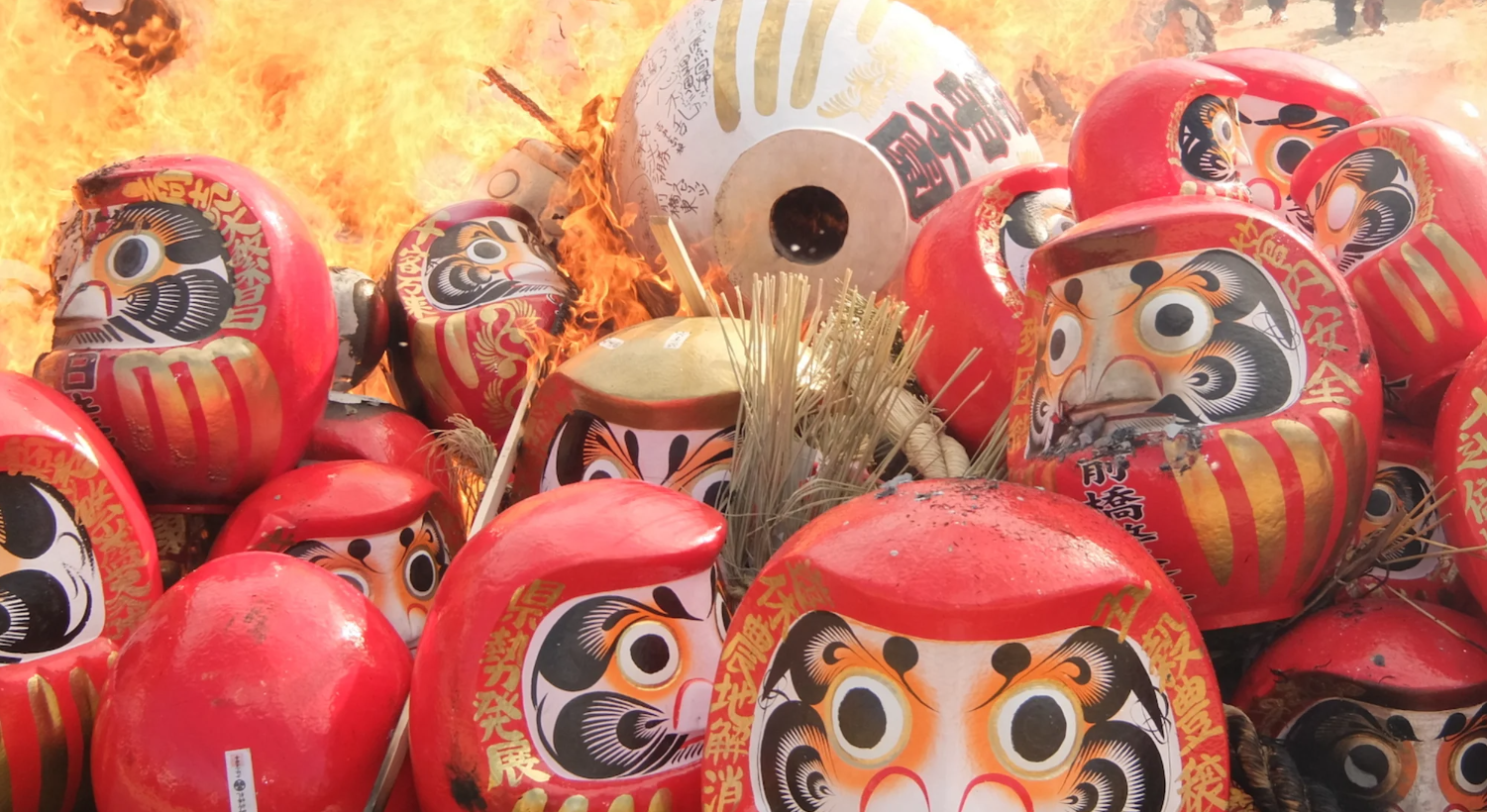 Several red and white large round Japanese daruma dolls with black circular eyes and a mustache lay in a pile among dried grass and are about to catch fire.