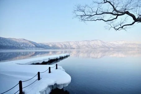 A winter view of Lake Towada showing a dock covered in snow and a cloudless sky reflected in the water.