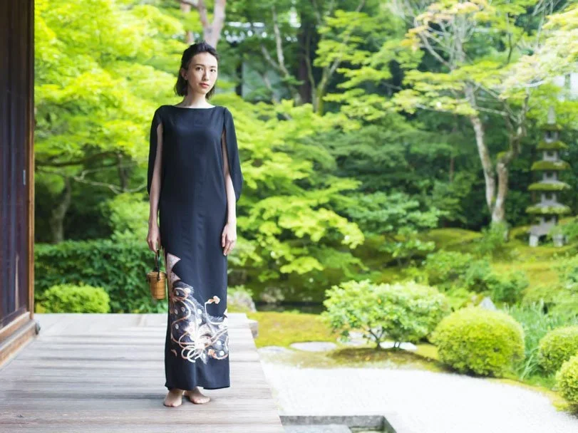 A woman stands in a Japanese garden wearing a long dress made out of a black formal Japanese kimono with hand painted rope stenciling on the bottom.