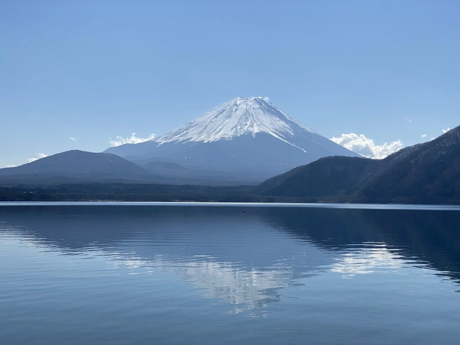 view of the foothills of and Mount Fuji with a lake and the reflection of the mountains in the foreground. Image is tinted blue with small low laying clouds on near the mountain on an otherwise cloudless sunny day .