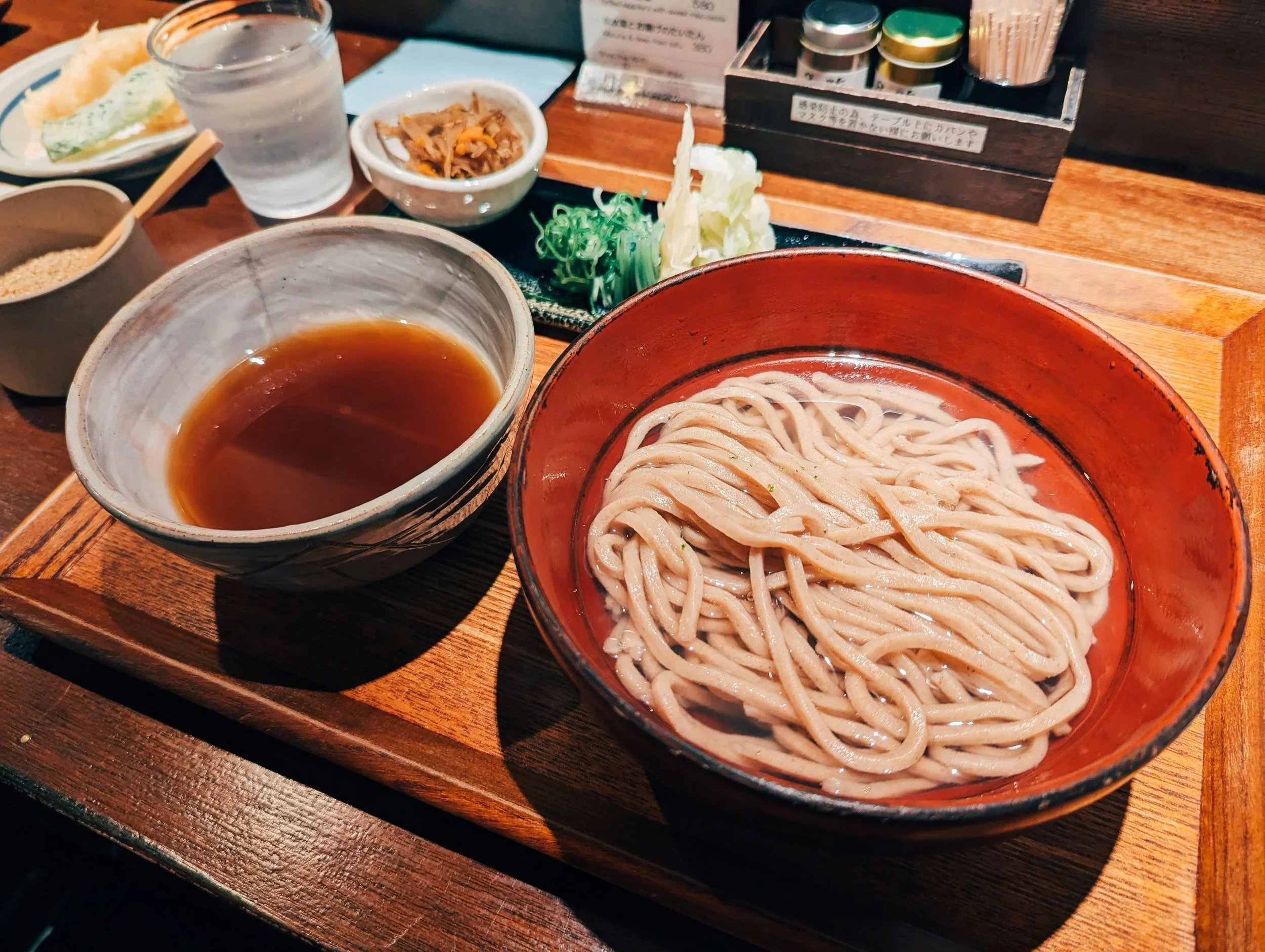 Image of a bowl of noodles with dipping sauce and vegetables on the side.