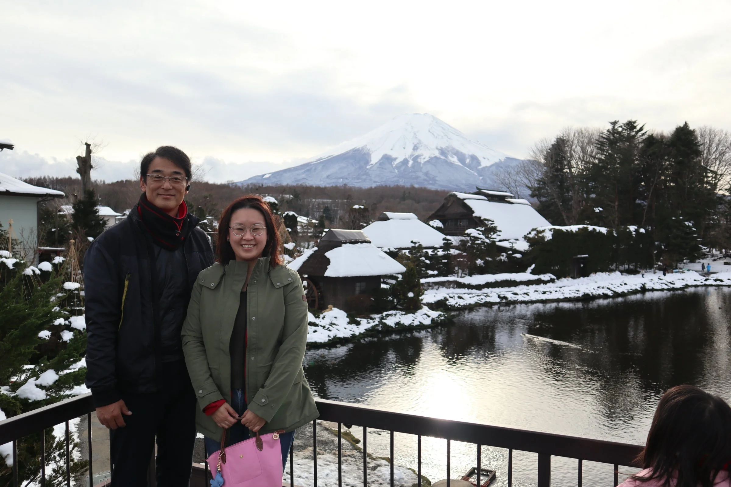 A male and female or Tokuda and Wanping, stand in front of a snow covered village in front of Mount Fuji.