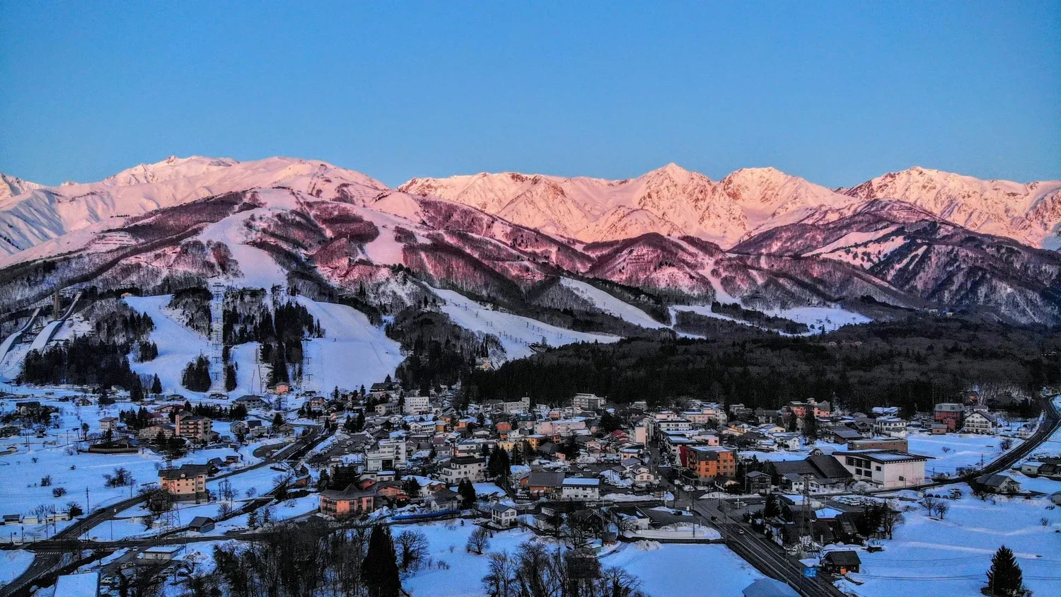 Snow-covered town at the base of snow-capped mountains during sunset.