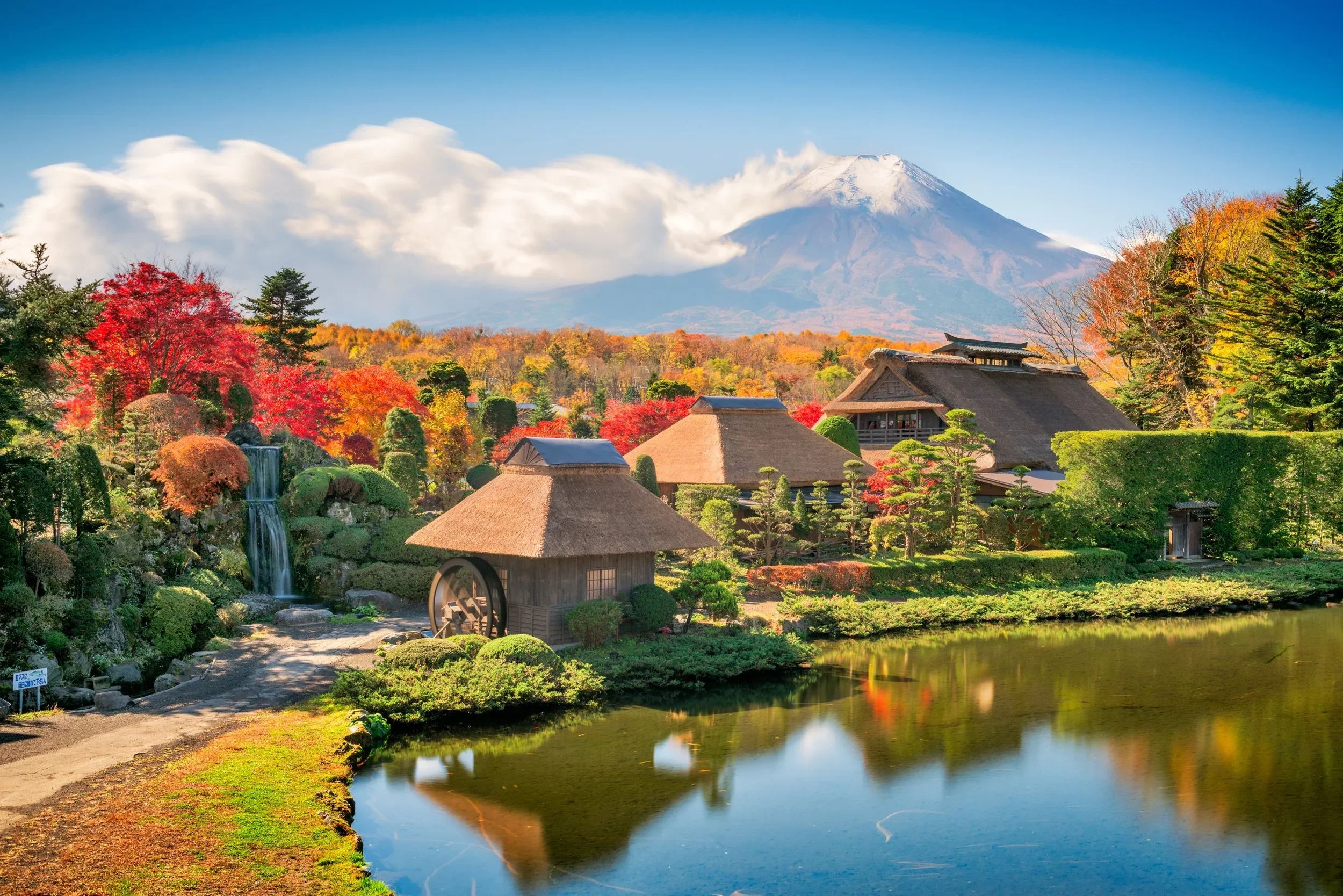 Small Japanese tatched roof huts in front of Mount Fuji on a mostly cloudless fall day, with only low-level clouds near the mountain and a pond in the foreground.