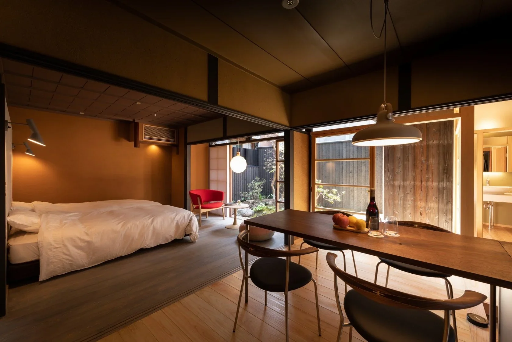 Japanese hotel room with 2 single beds, a seating area with a red chair and a kitchen table in the foreground. View from the corner of the room looking out to a small Japanese garden.