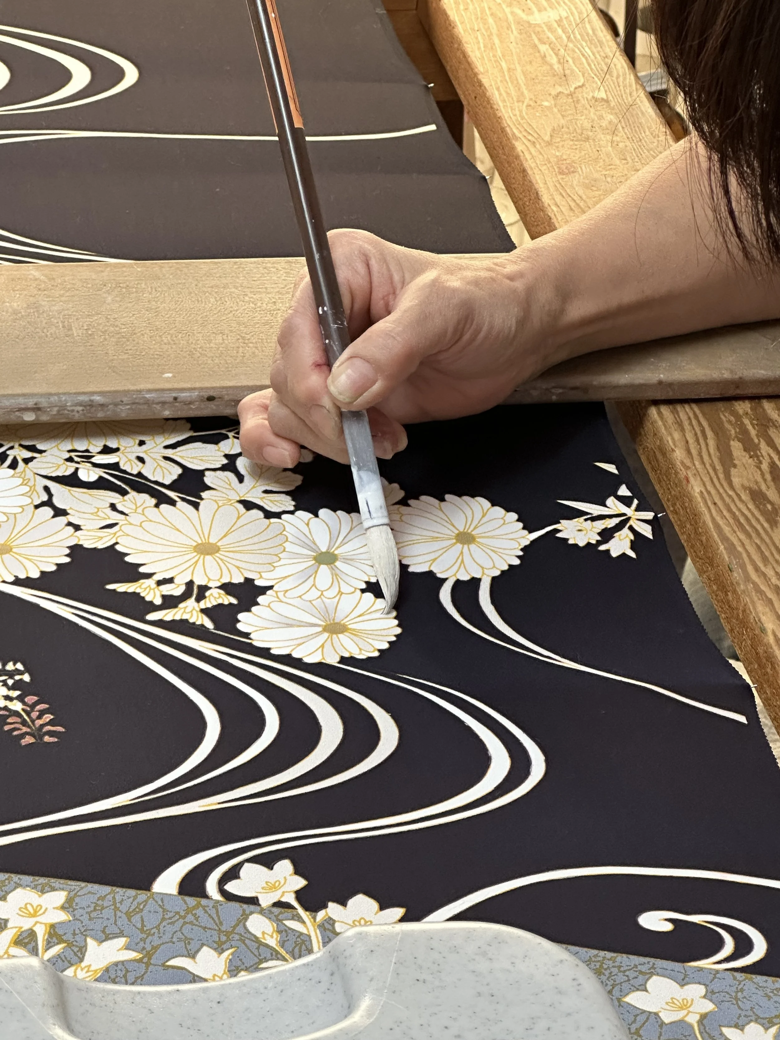 Closeup of a textile Yuzuen painter paints white paint on a kimono dyed black. Image of type of factory visited on tour.