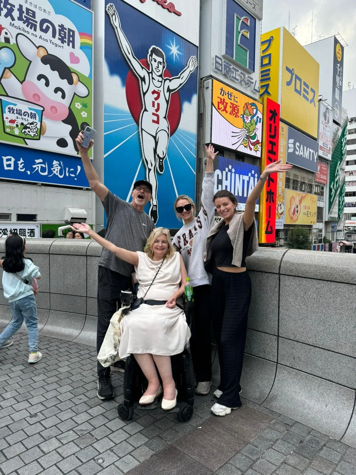 Four people, including a woman in a wheelchair, posing and smiling with their arms raised in front of colorful Japanese billboards in an urban area. One person is holding a phone, and a child walks past on the sidewalk.