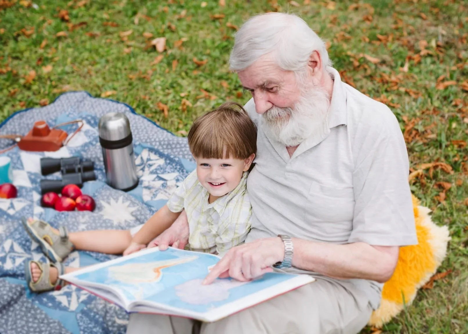 Older man reads to a child sitting in their lap on a blanket on top of the ground.