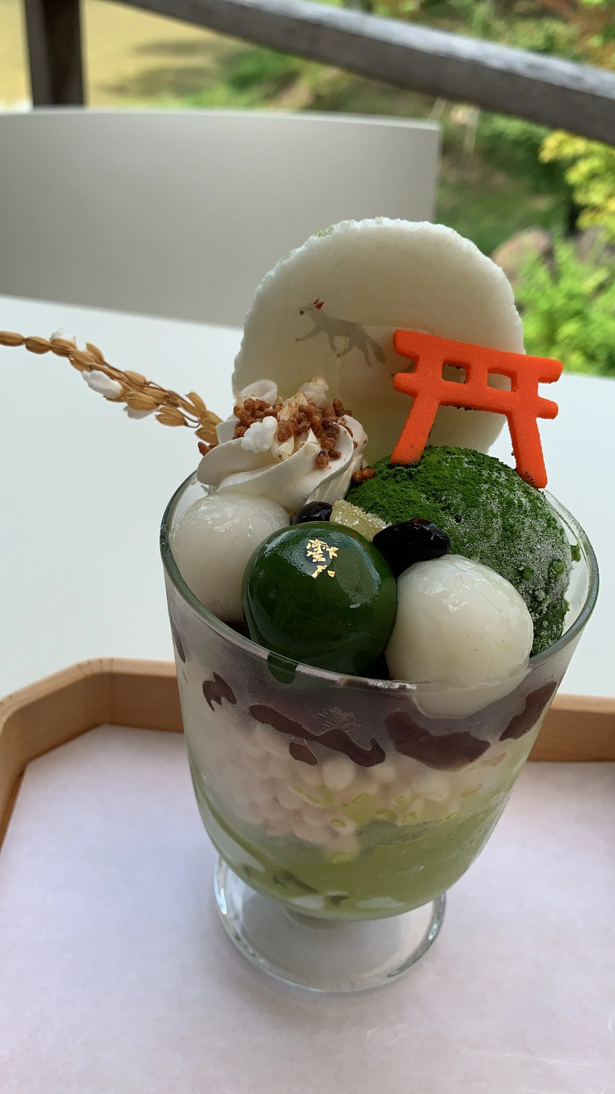 A matcha parfait with rice crackers and dango with ice cream a fox senbei and a red tori gate in a small cup resting on a wooden tray on a table in a Japanese garden in Fushimi Inari Shrine in Kyoto, Japan.