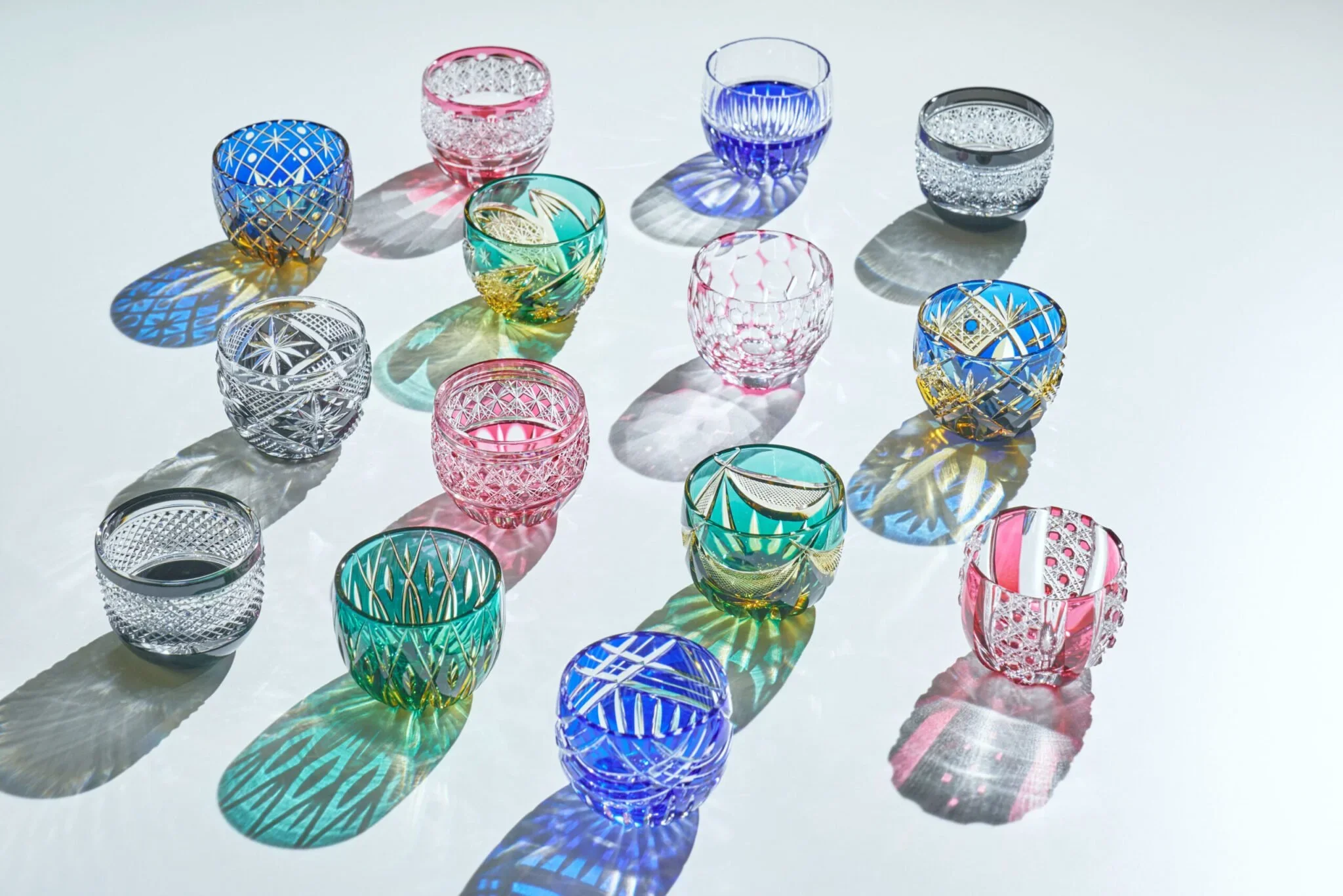14 different and colorful etched glass mini cups for sake are arranged on a white background with harsh light to showcase the etched glass patterns.