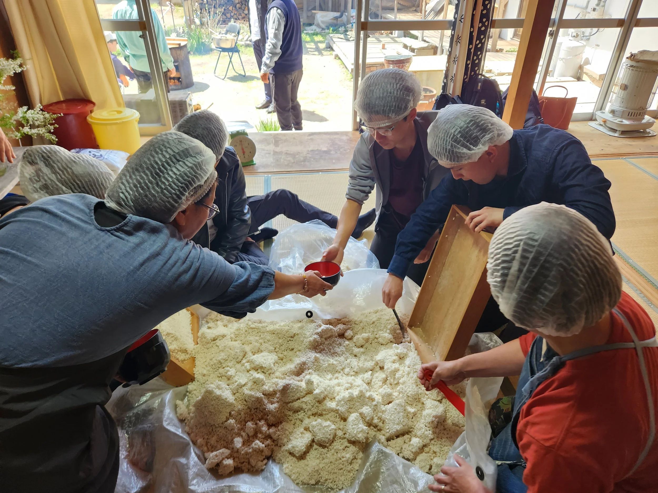 A group gathers inside an old Japanese farm house to make miso in the traditional style Japanese families made miso, a woman passes the bowl to a younger person.