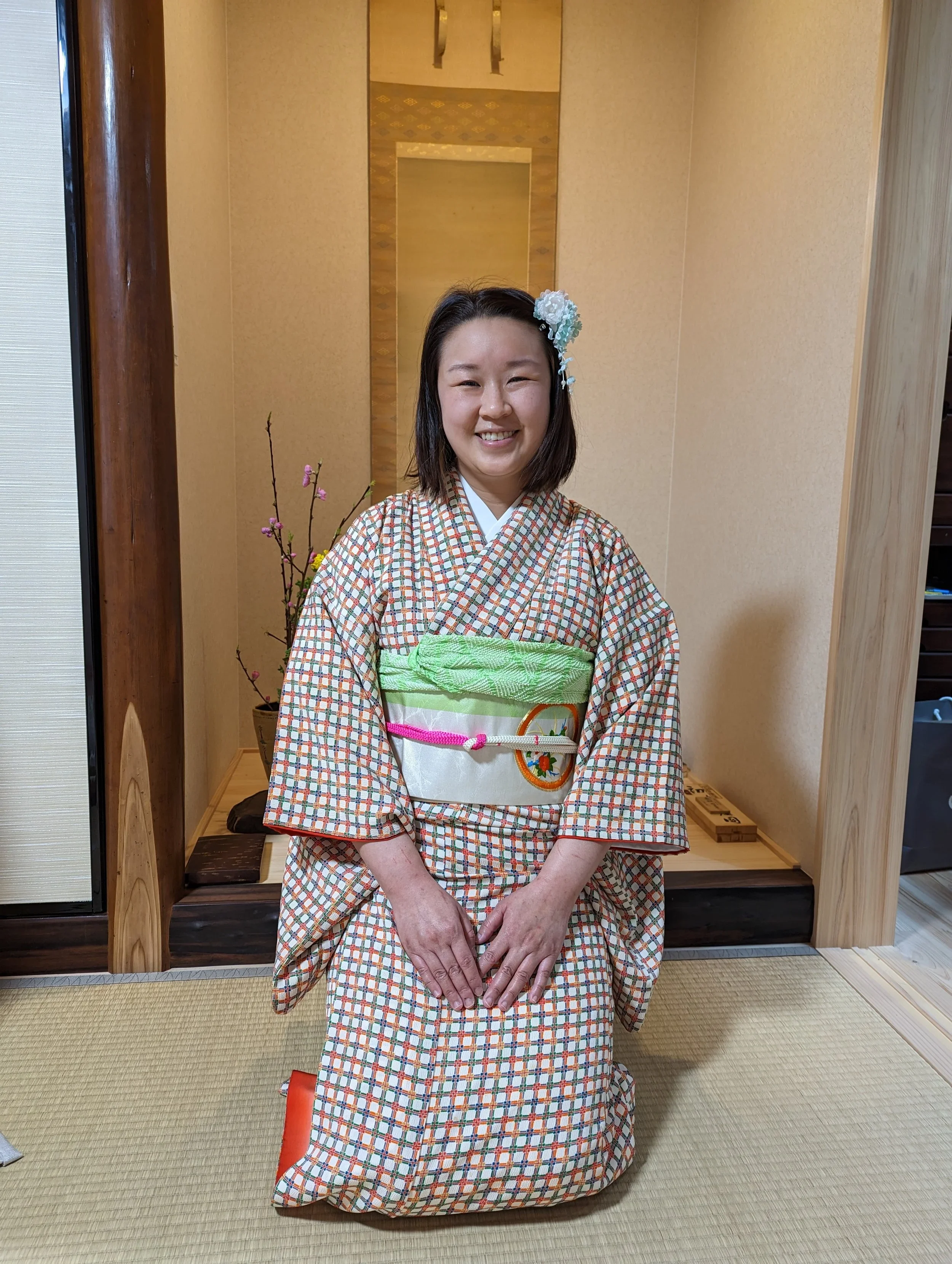 Young asian woman is seated in a checkered kimono with a white and green obi or sash in front of a gold Japanese painting.
