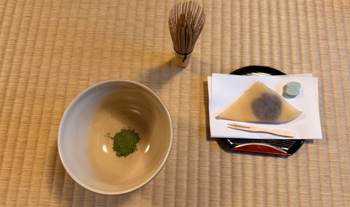 Powdered matcha in a bowl with a Japanese sweet placed next to it and a bamboo whisk placed upside down in front on a tatami mat floor.
