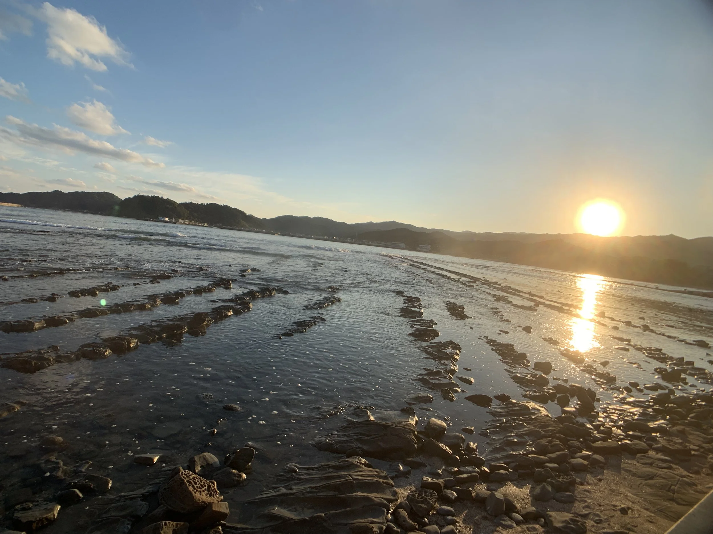 Sunset over the ocean with hills in the background, shoreline with rocks and wet sand in the foreground.