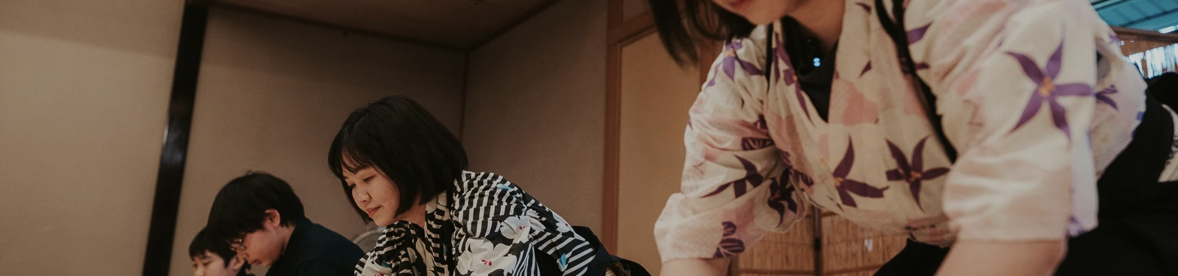 Several people are seated in a row in a japanese room wearing kimono and are bowing their heads.