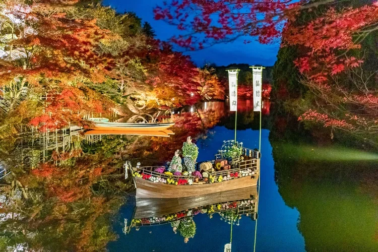 A traditional wooden Japanese boat floats in a lake decorated with with fall flowers and two tall flags, surrounding the lake are fall colors and a mountain. Scene is at dusk. Crysanthemum and Momiji Festival at Hirosaki Park.