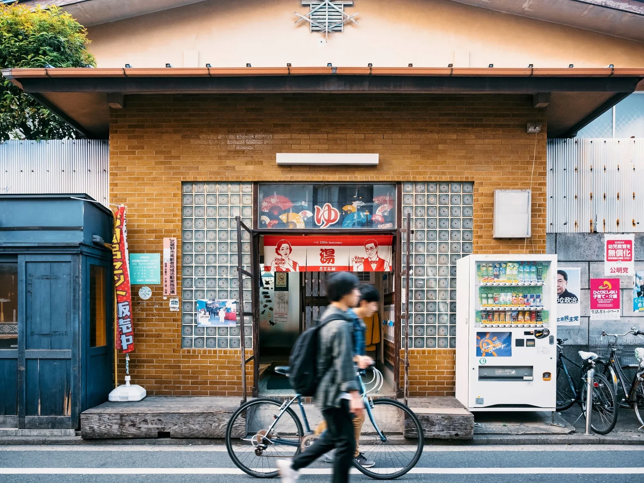 The exterior of an orange brick sento, or Japanese bath house, in a urban street scene with a bicyle, traveler, and vending machine out front.