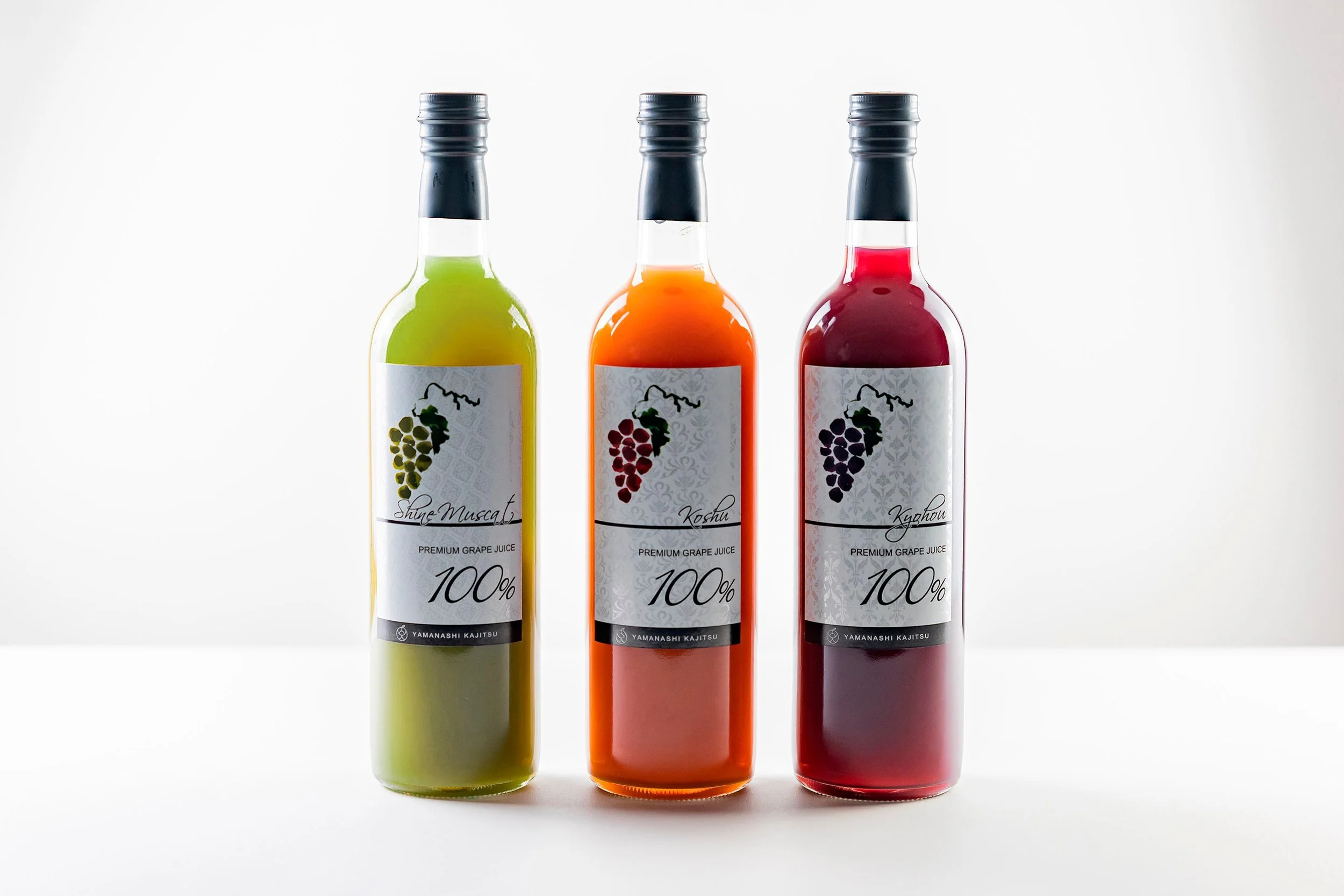 Three bottles of green, orange, and red grape wines in a line on a white table with a white backdrop.