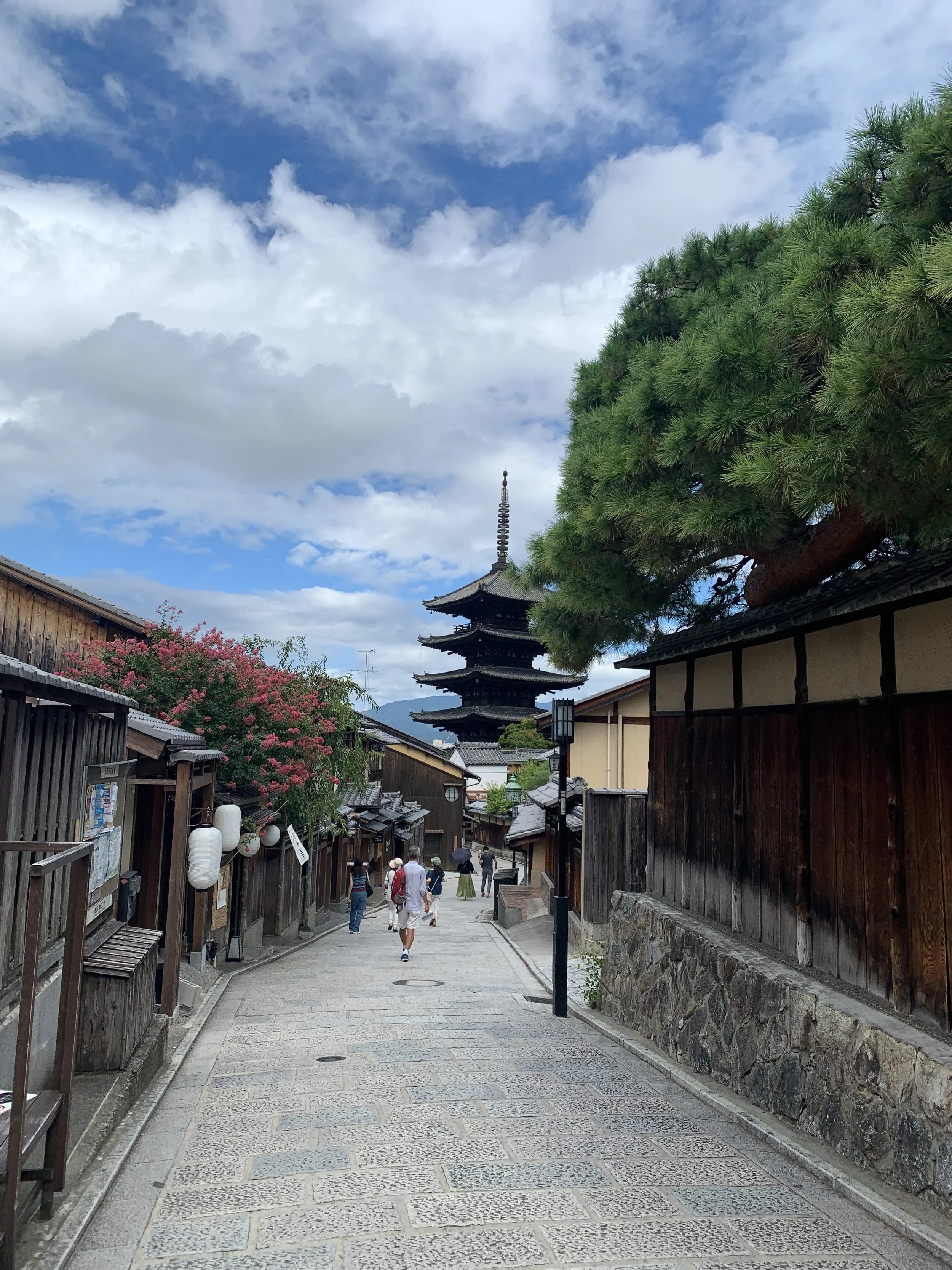 A narrow street in Japan with wooden fencing and short wooden buildings with a stone road and a 5 story wooden pagoda at the end on a sunny cloudy day in Kyoto, Japan.