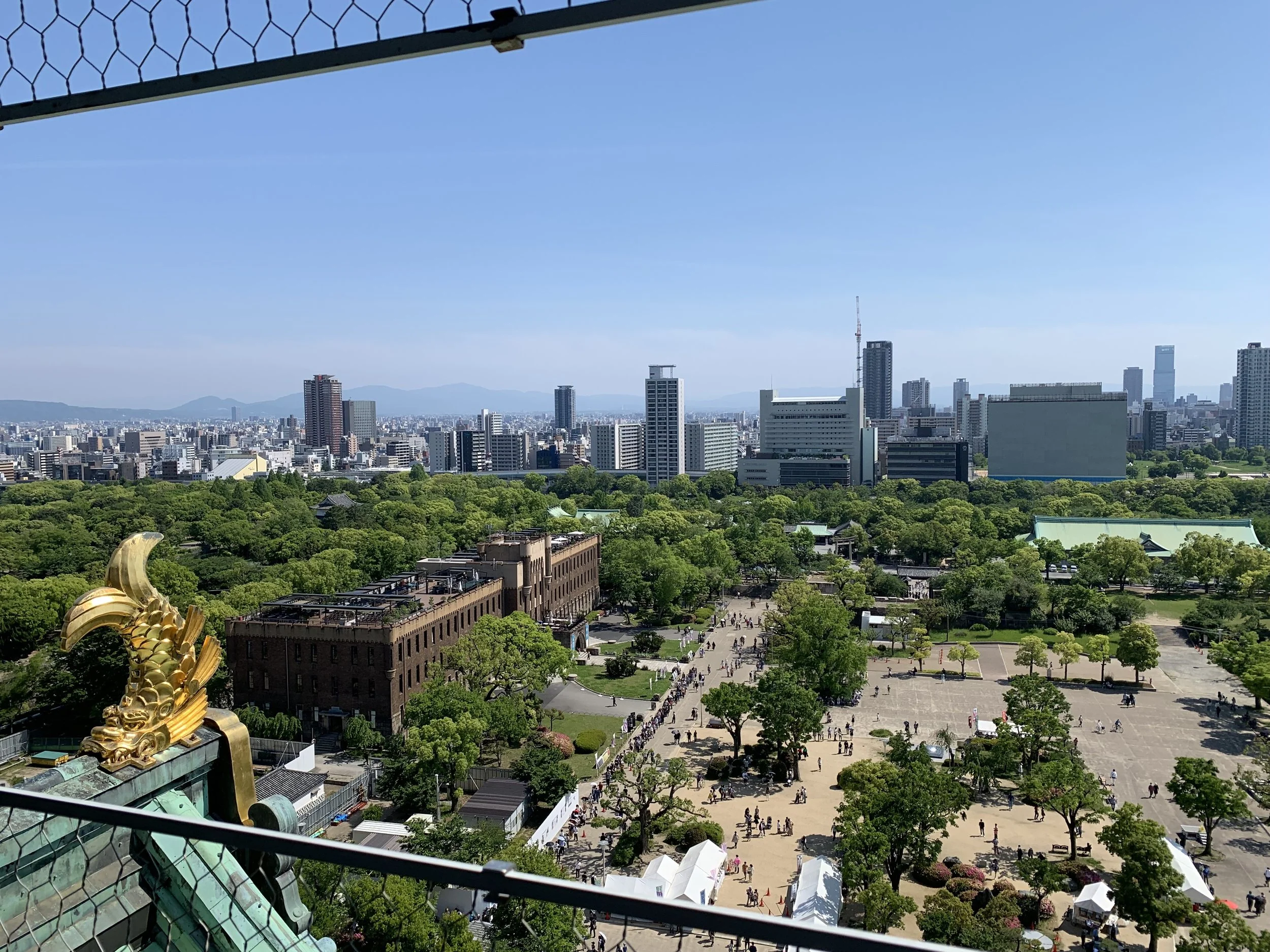 View from a top floor of Osaka castle looking out thorough a portal, with a partial view of the gold roof ornament, a view of the park and people below and city buildings and the distant mountains on a clear blue sky day.