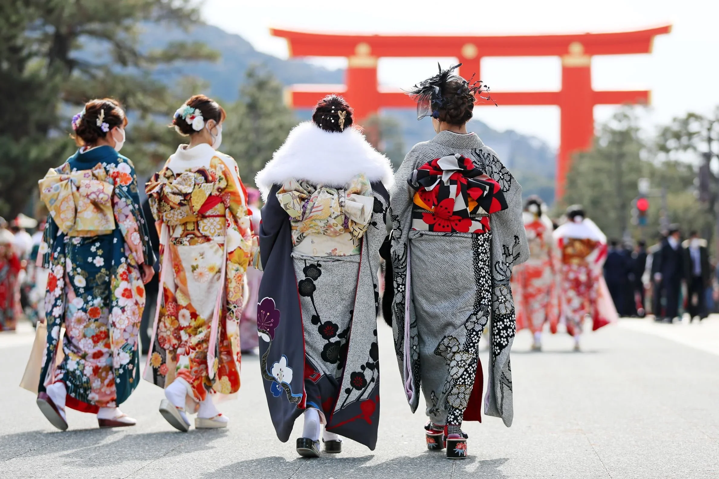 4 ladies dress in a winter furisode kimono of many colors including balck and red and oranges, one with a white fur scarf walking towards a tall Japanese torii gate.