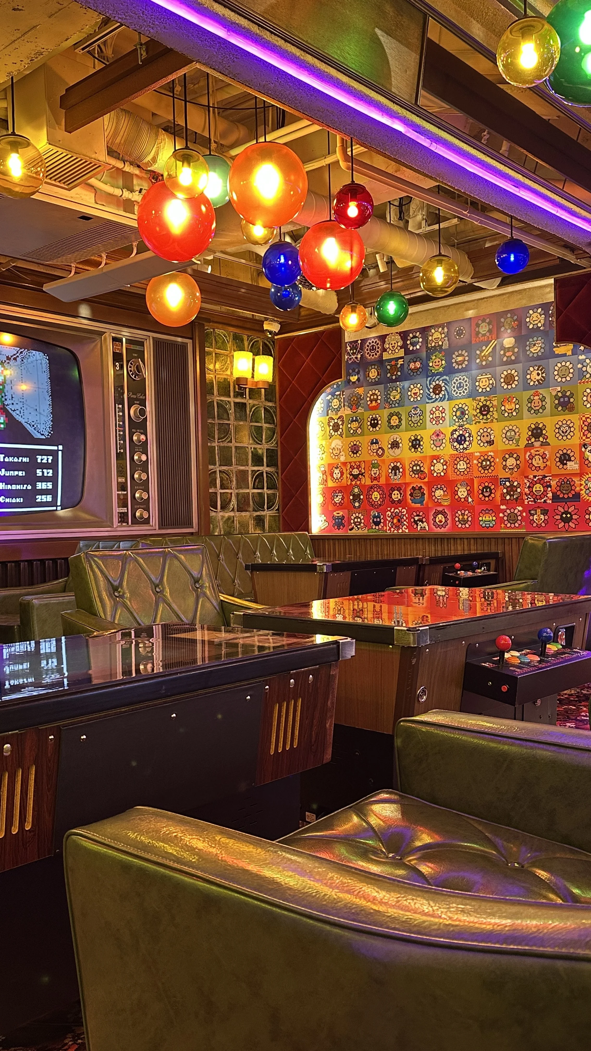 A retro arcade game room decorated with colorful pendant lights hanging from the ceiling, a vintage television, and framed pixel art on the wall. There are green leather chairs and a pinball machine in the foreground.
