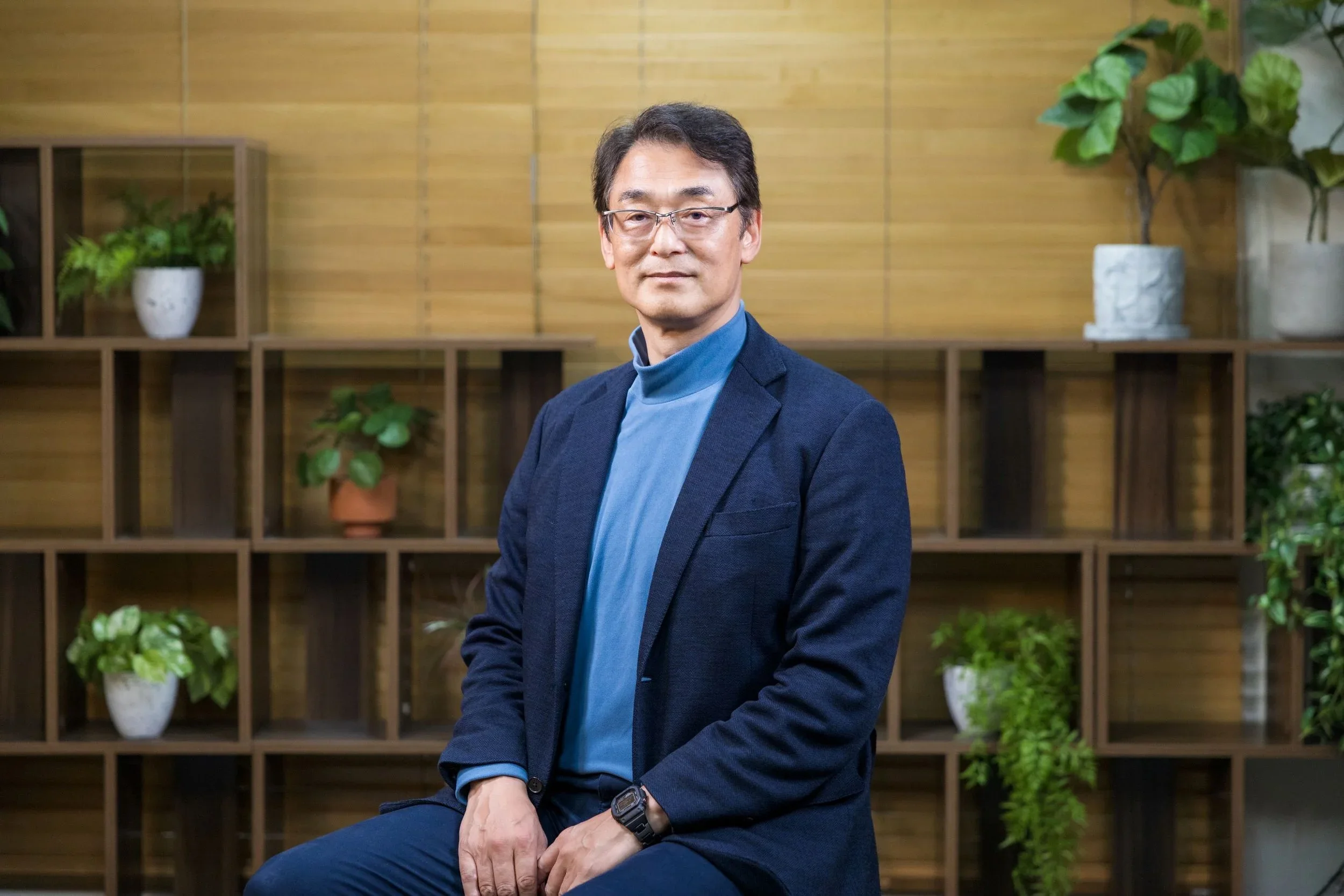A man wearing glasses, a blue turtleneck, and a dark blazer, seated in front of a wooden bookshelf with potted plants, looking at the camera.