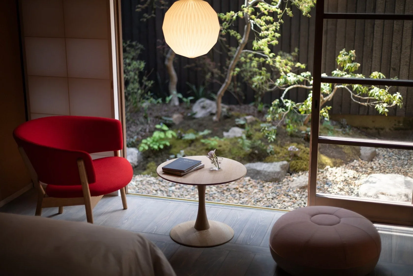A seating area  with a cushion stool, round wooden table and a red chair with a warm origami oblong lamp in a hotel room overlooking a small Japanese garden.