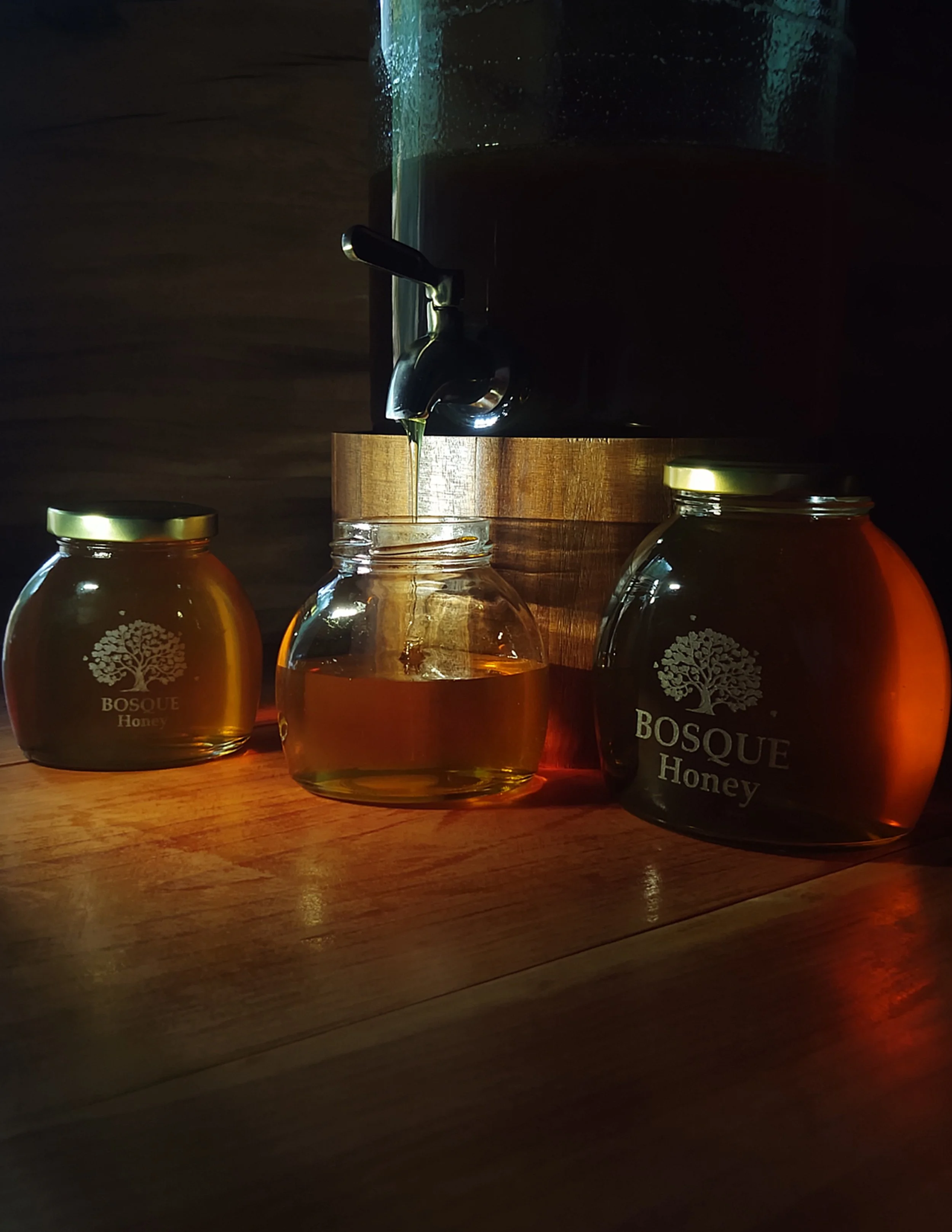 Three jars of honey labeled 'Bosque Honey' on a wooden surface, with a tank of honey above, pouring honey into one jar.