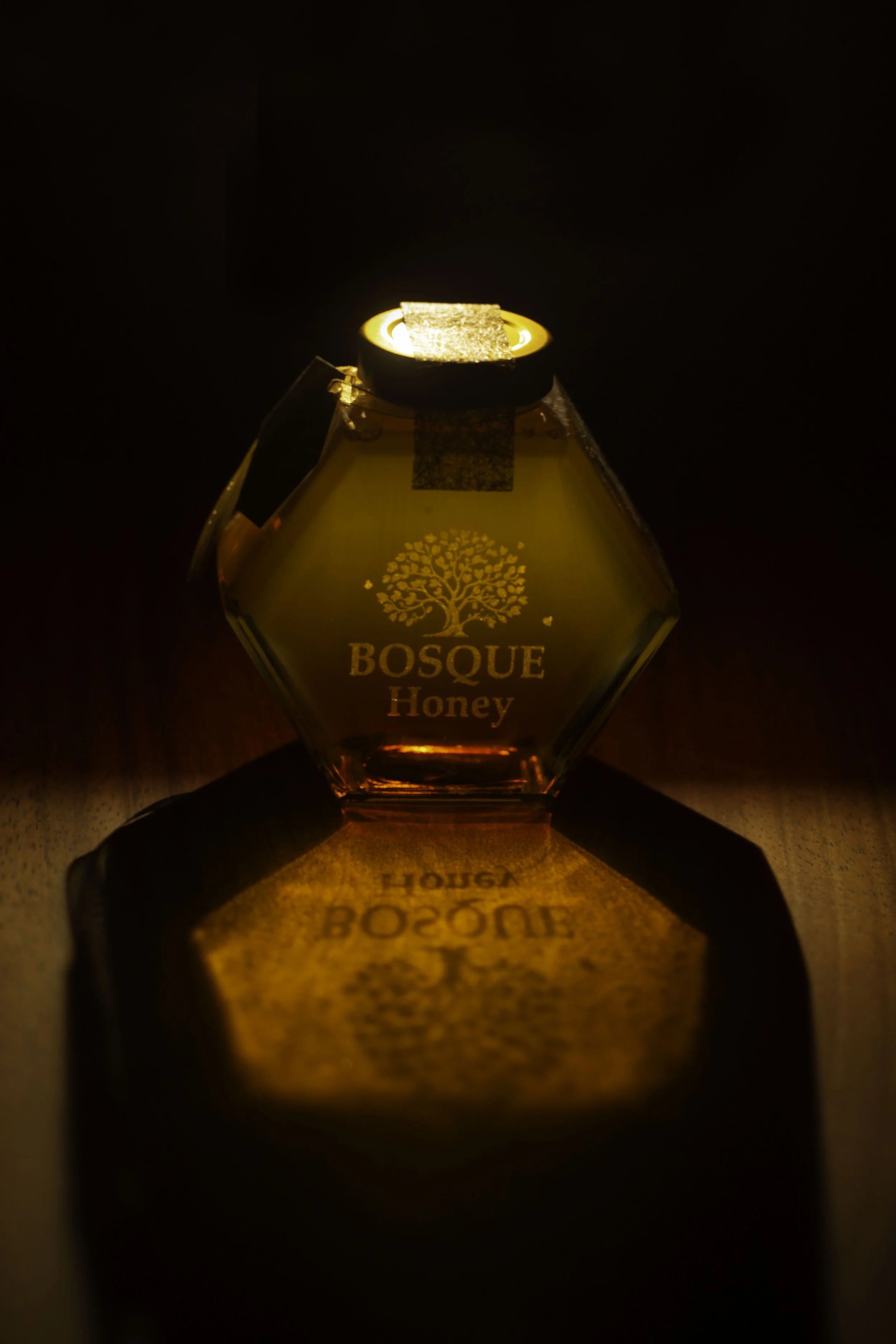 A jar of Bosque Honey illuminated by a warm light, reflecting on the surface below.