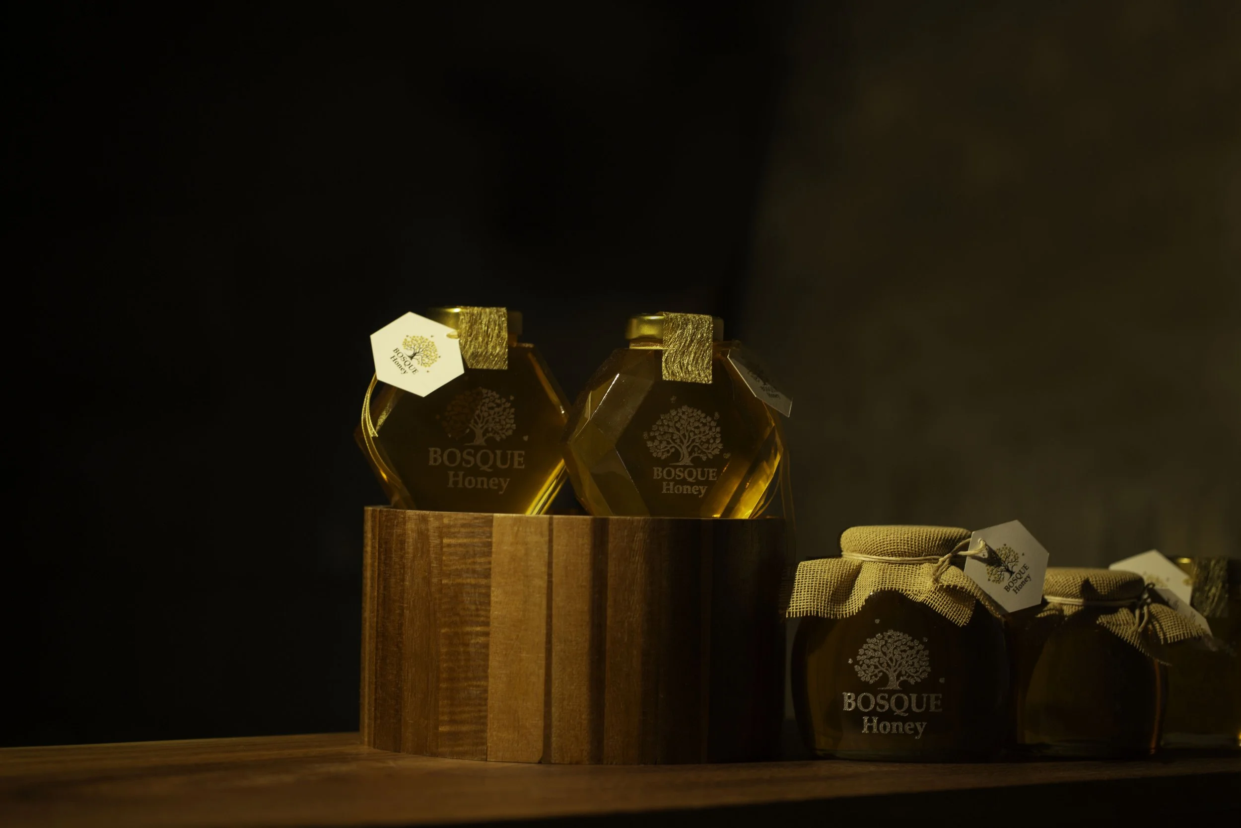 Two jars of Bosque Honey placed on a wooden surface with tags, and additional honey jars with tags and a cloth underneath, all set against a dark background.