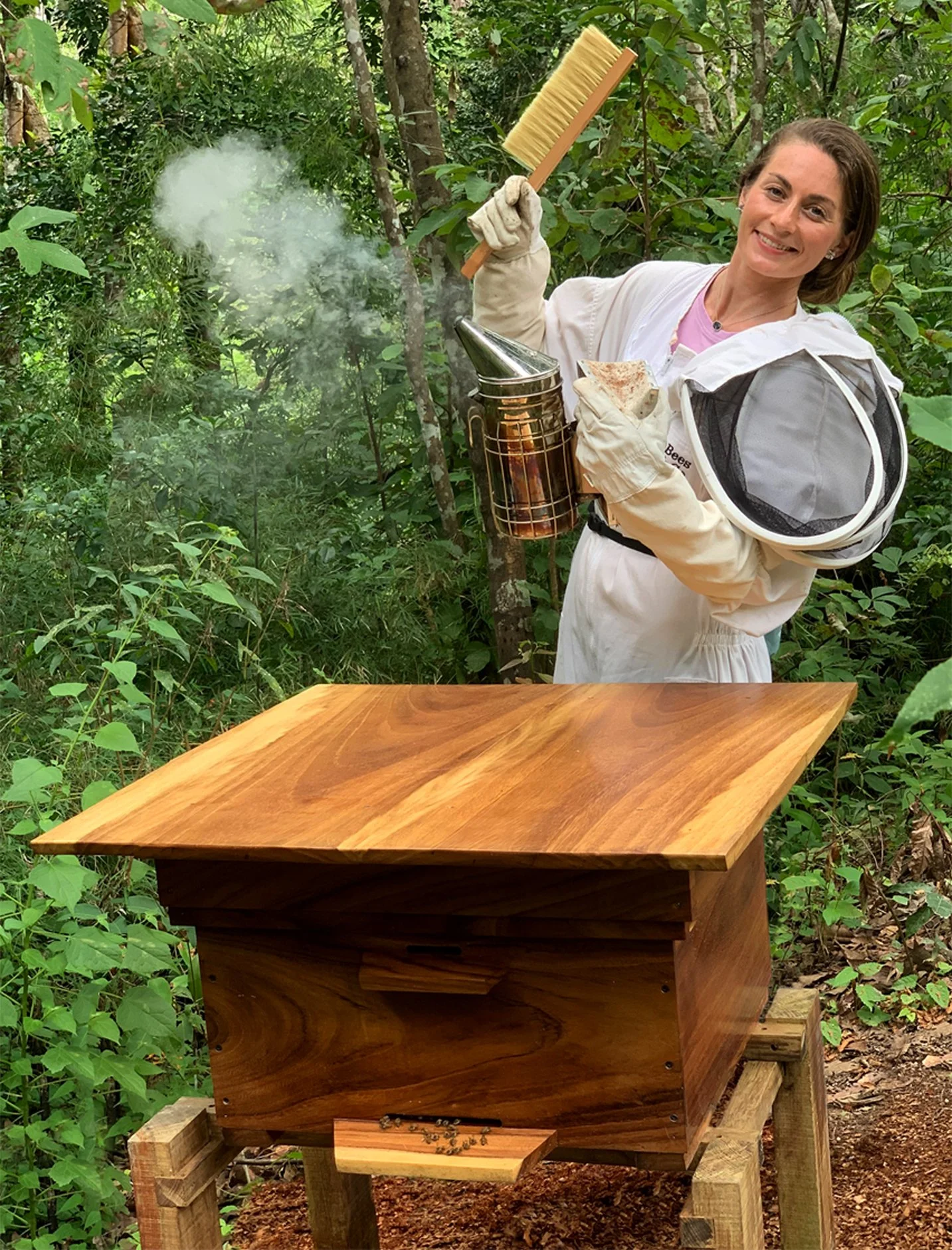 A woman in a white jacket and gloves holding a bee smoker and a piece of honeycomb, standing next to a beehive made out of Parota wood of the same forest area in a wild mountain area.