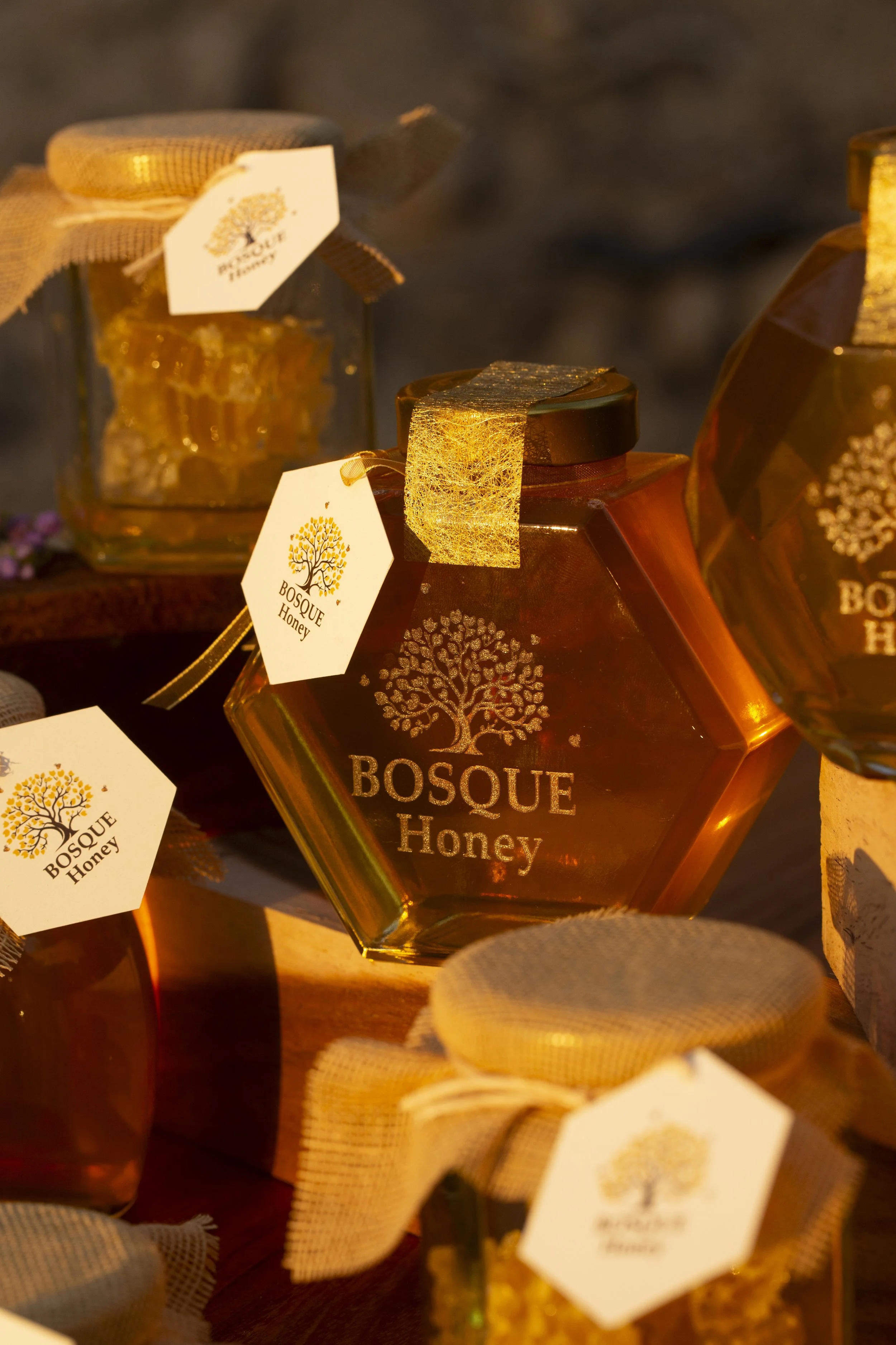 A honey jar labeled 'Bosque Honey' with a tree logo, golden ribbon, and a gold decorative element on the lid. Several other honey jars are visible in the background.