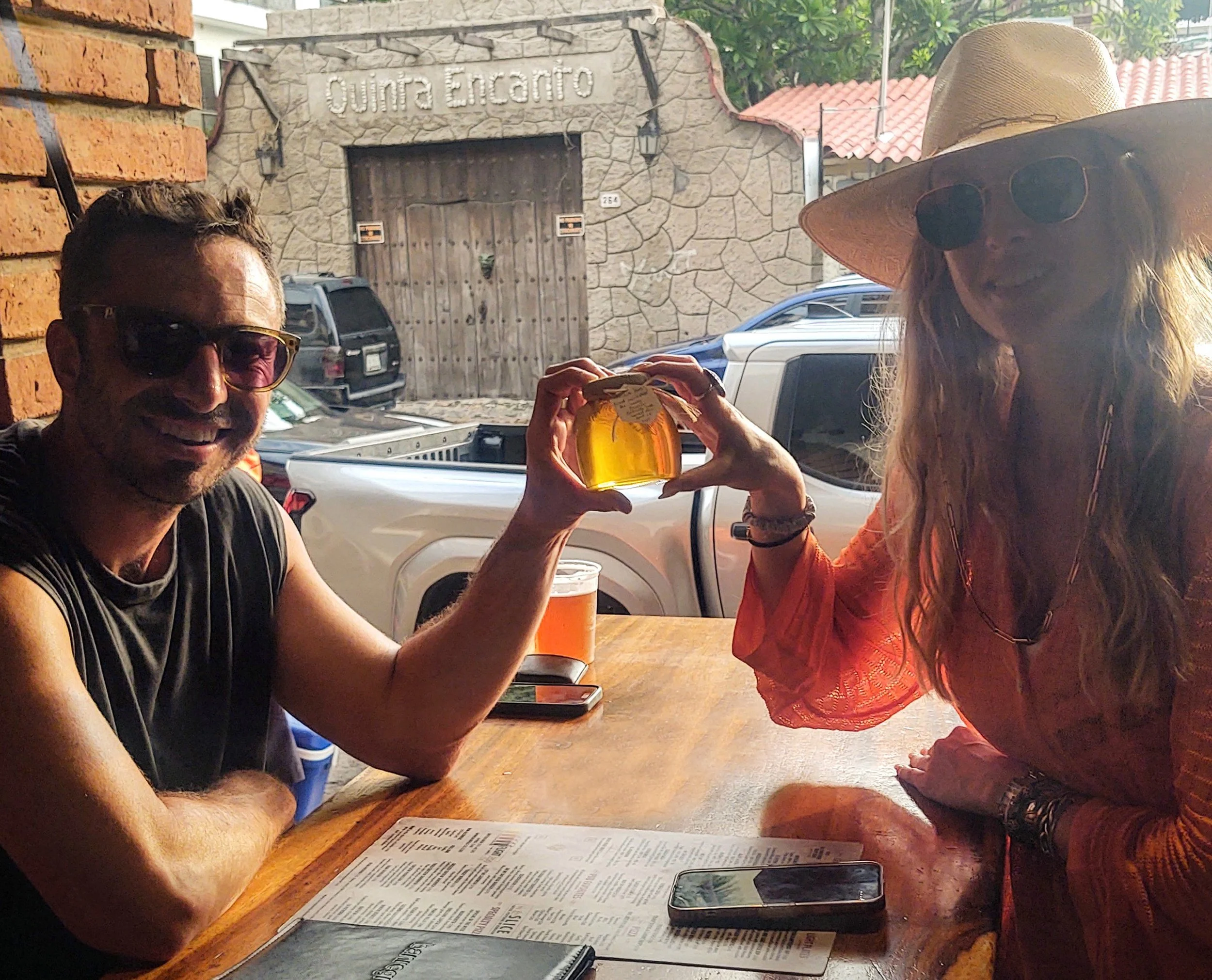 Two people sitting at a wooden table in an outdoor setting, holding a glass of beer together. The man on the left is smiling and wearing sunglasses, and the woman on the right is wearing a large straw hat and sunglasses. There are cars parked in the background, and a stone building with a wooden door that has a sign reading 'Quinta Encanto.'