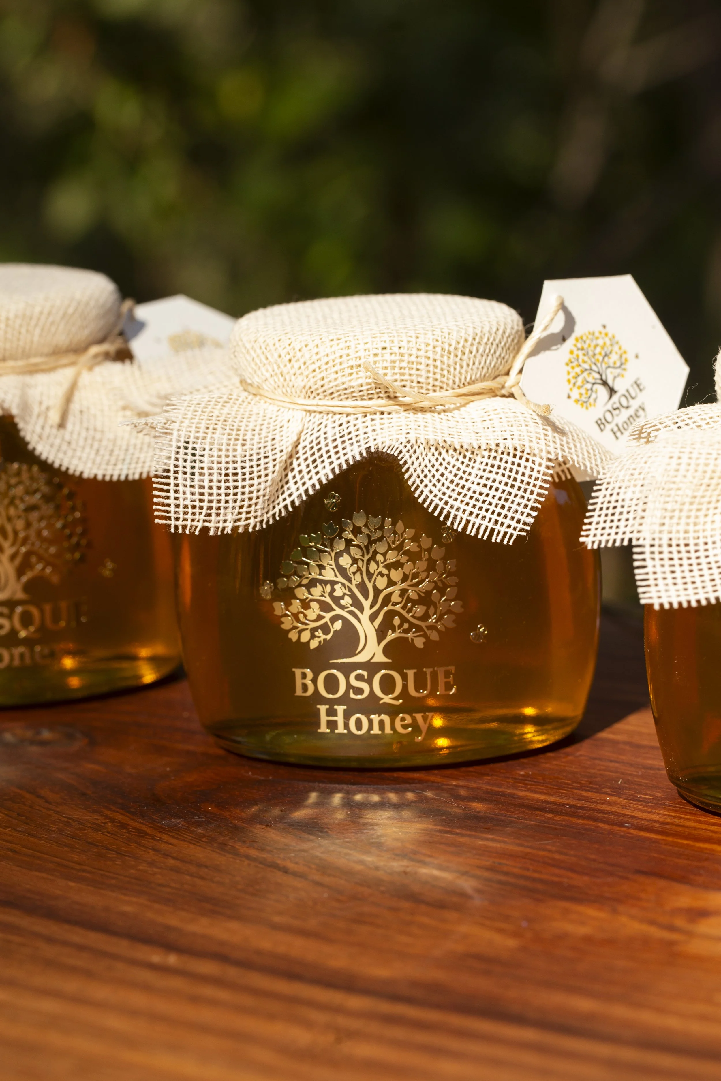 Glass jars of honey with beige fabric covers and tags that read "Bosque Honey" on a wooden surface, with blurred background.
