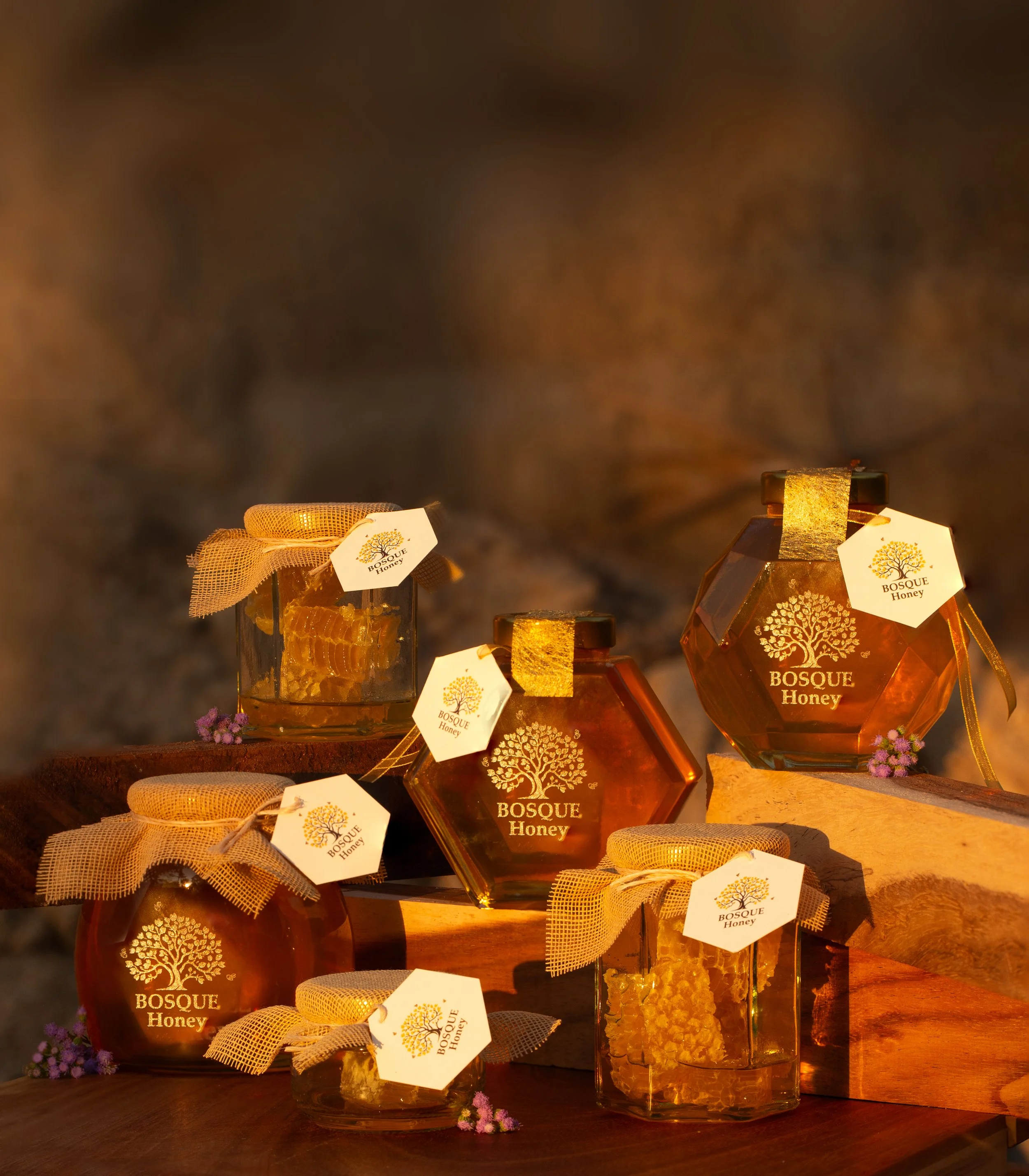 Various jars of honey labeled 'Bosque Honey' with tags, ribbons, and decorative elements displayed on a wooden surface, illuminated with warm lighting.