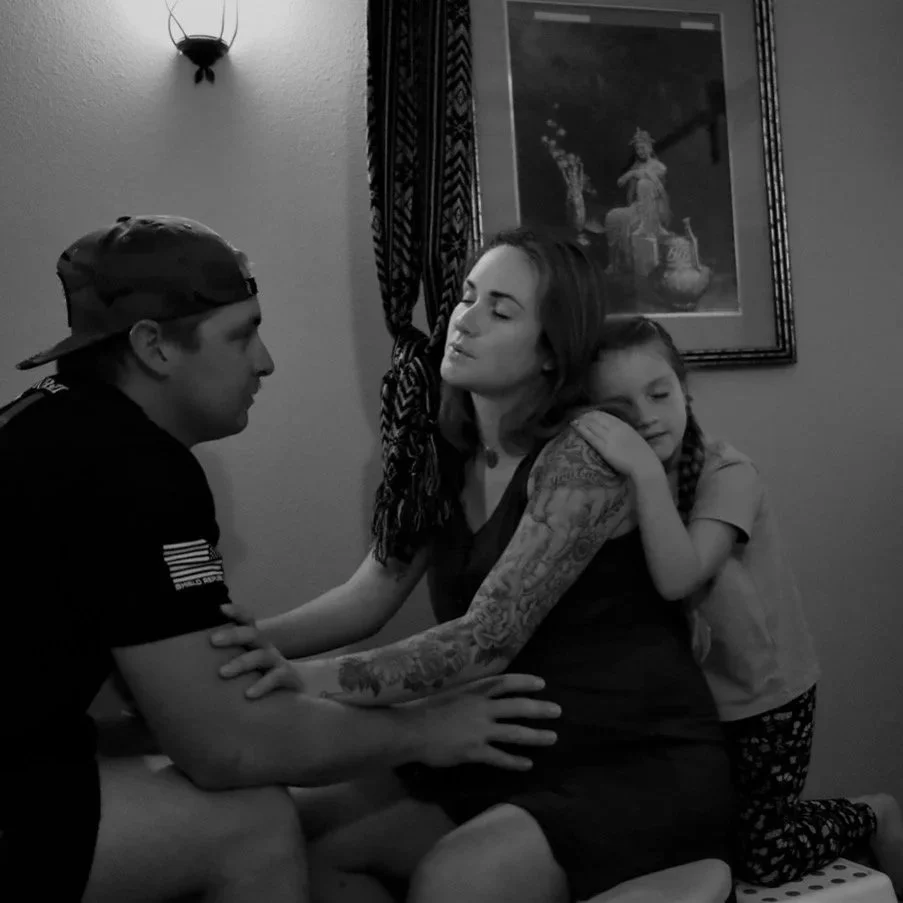 Family moment with three people: a man and a woman sitting close and a young girl hugging the woman from the side, in a cozy room with framed artwork and a wall sconce.
