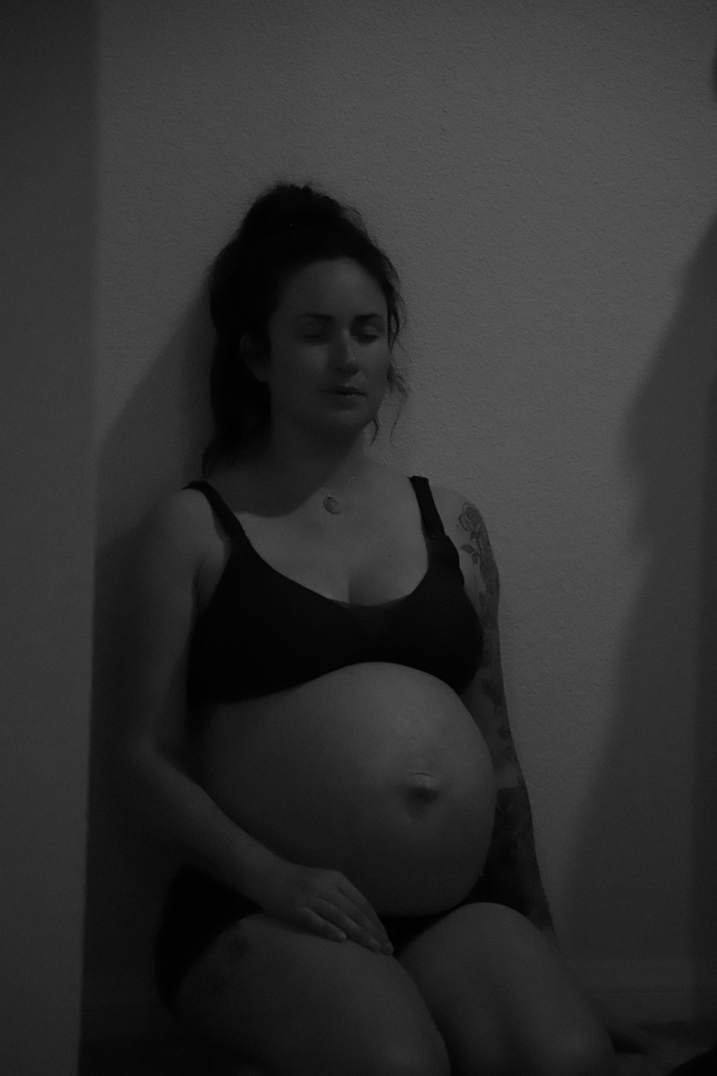 A pregnant woman with dark hair tied up stands against a plain wall, wearing a black sports bra and shorts, with tattoos on her arm.